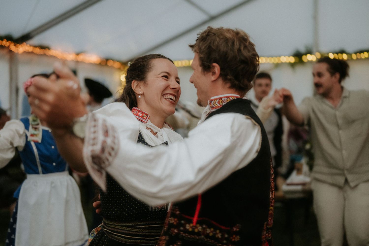 Couple dancing joyfully in traditional folk costumes at a festive cultural celebration under a decorated tent.