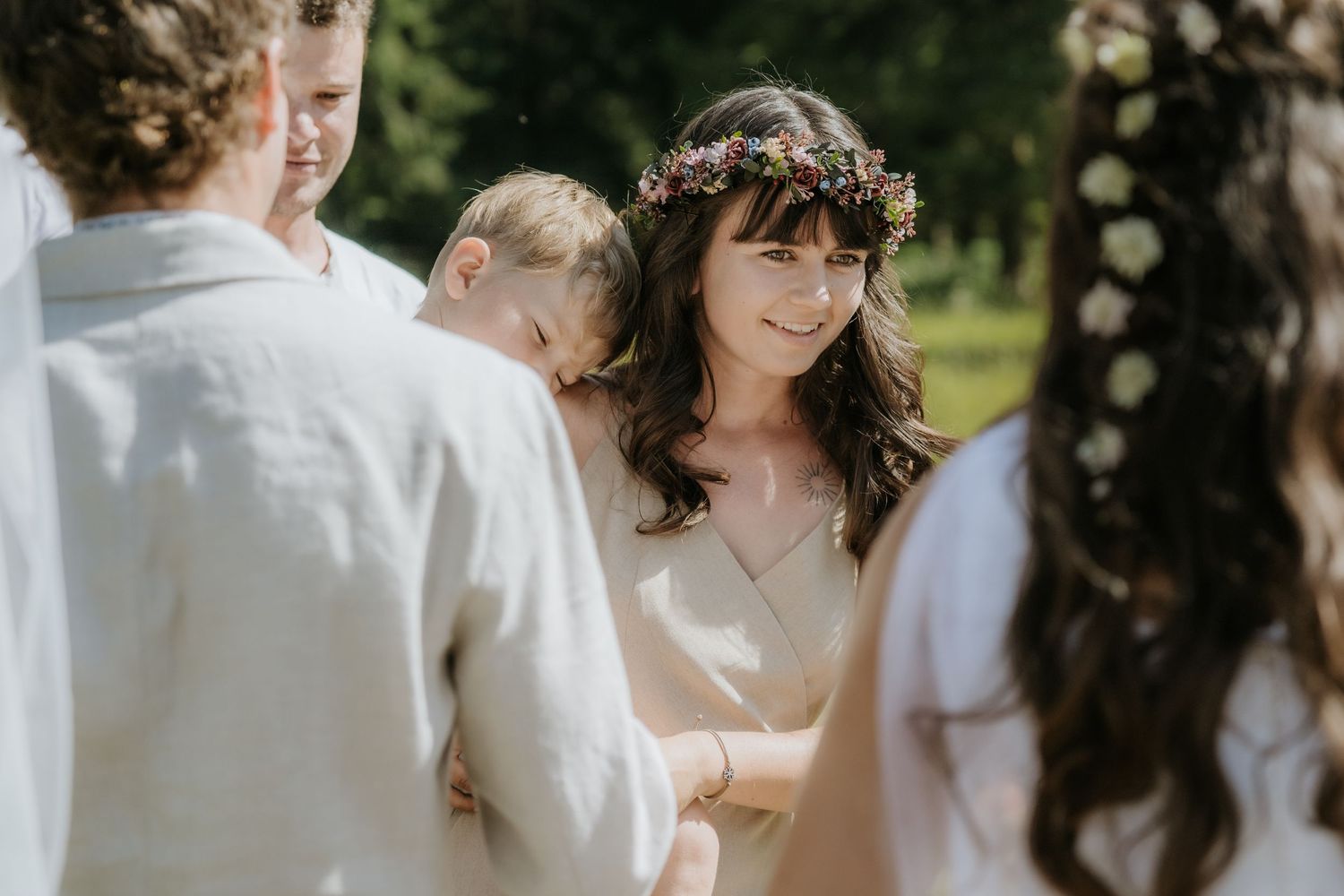 Woman wearing floral crown smiling during outdoor wedding ceremony surrounded by guests in natural sunlight.