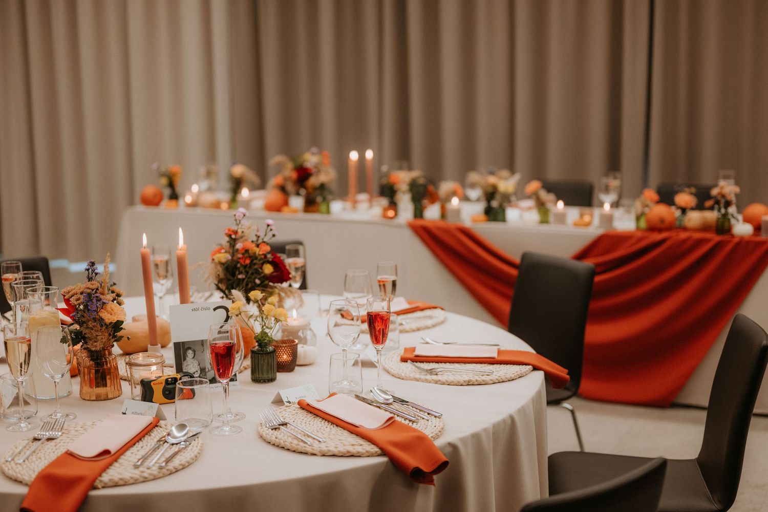 Elegant fall wedding reception table with orange napkins, candles, and colorful floral centerpieces on white tablecloths.