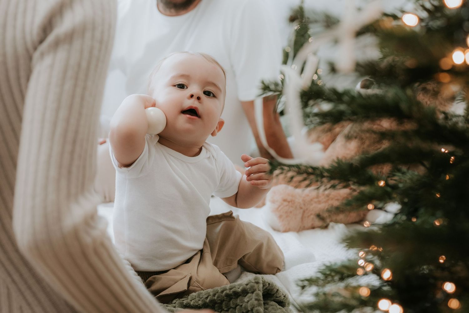 Curious baby in white onesie reaches out near a decorated Christmas tree with warm bokeh lights.