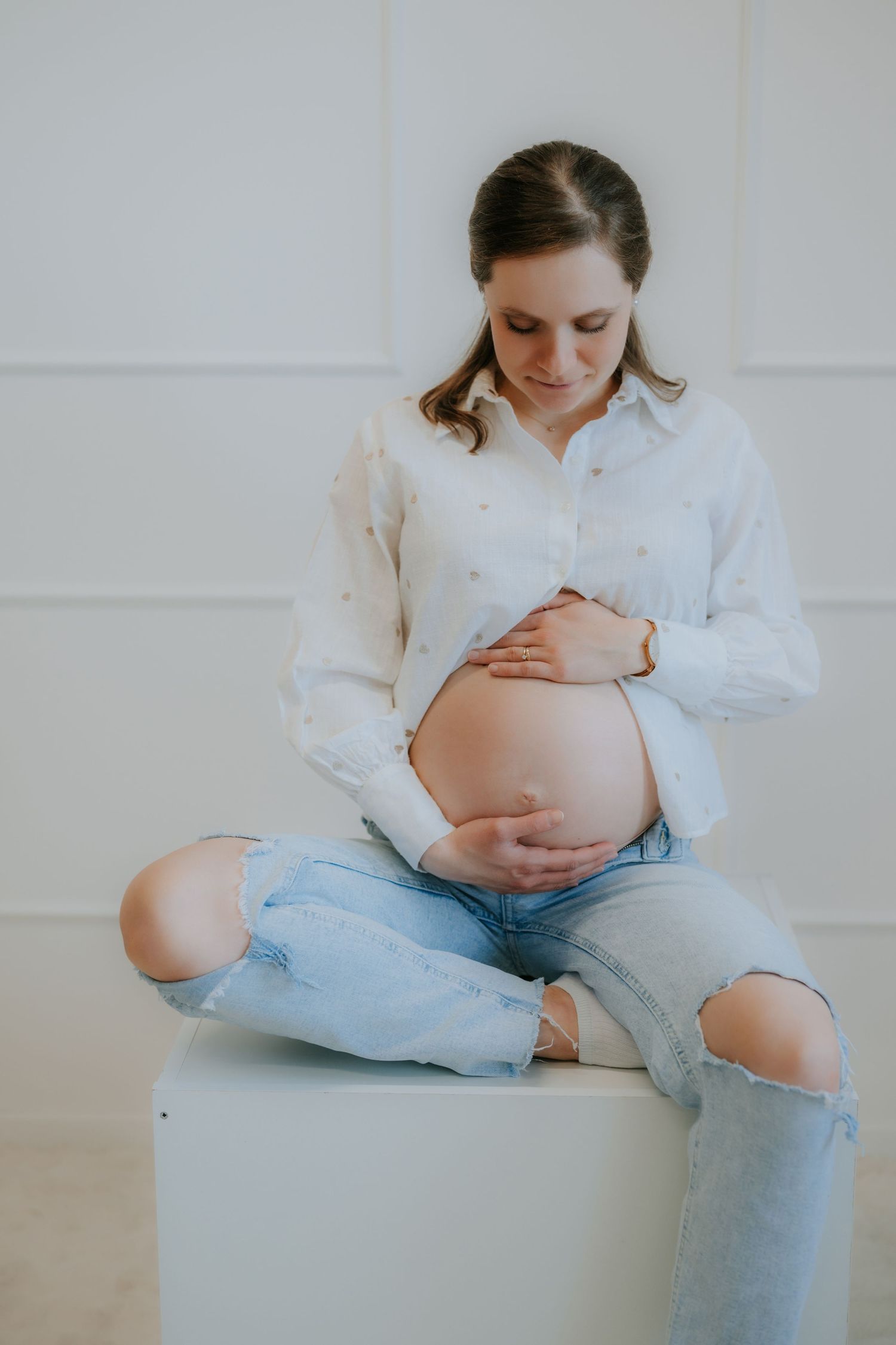 Pregnant woman in white shirt and ripped jeans sitting cross-legged cradling her baby bump.