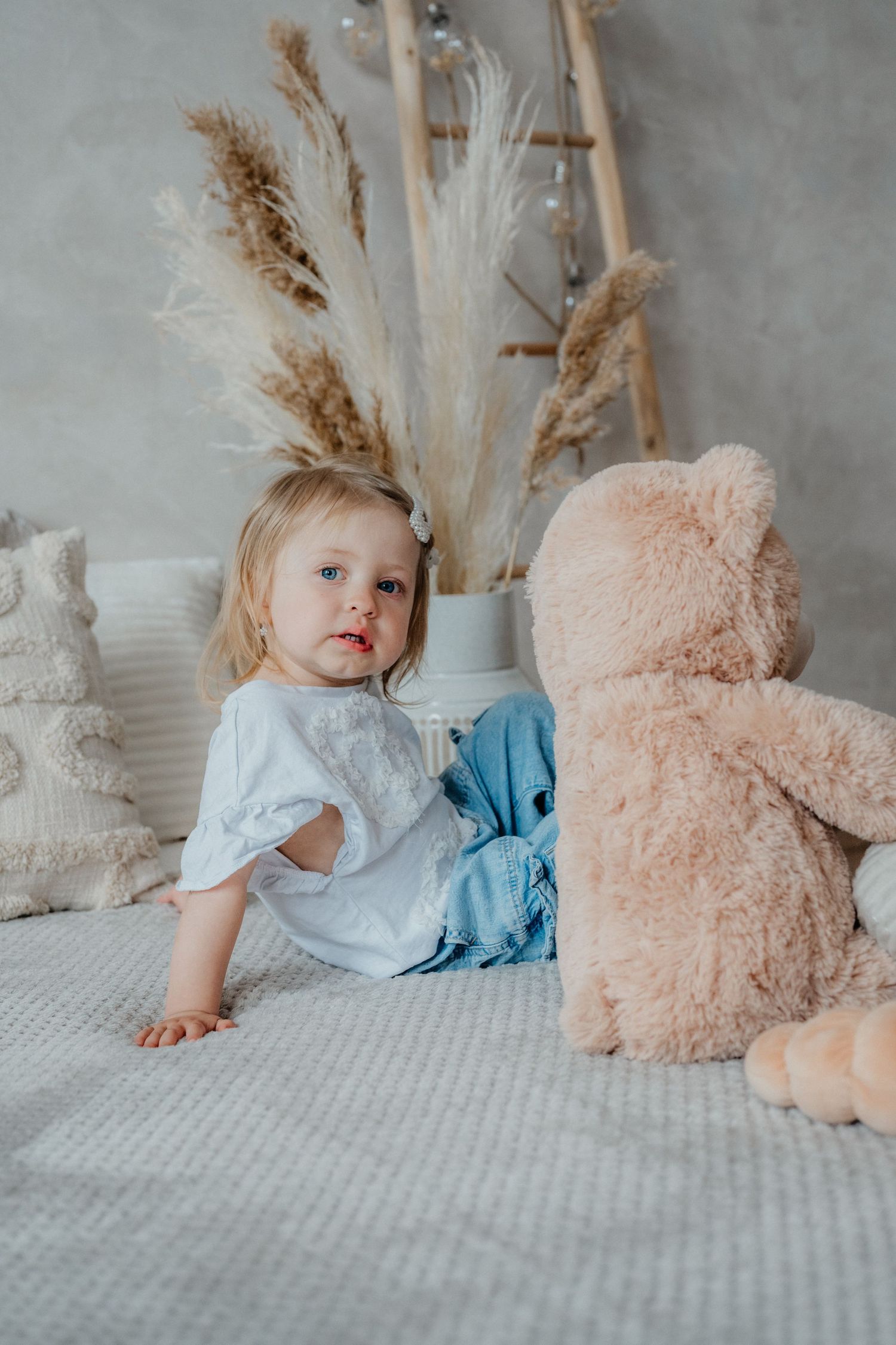 Blonde toddler girl in white top sitting on bed with large teddy bear and boho decor.