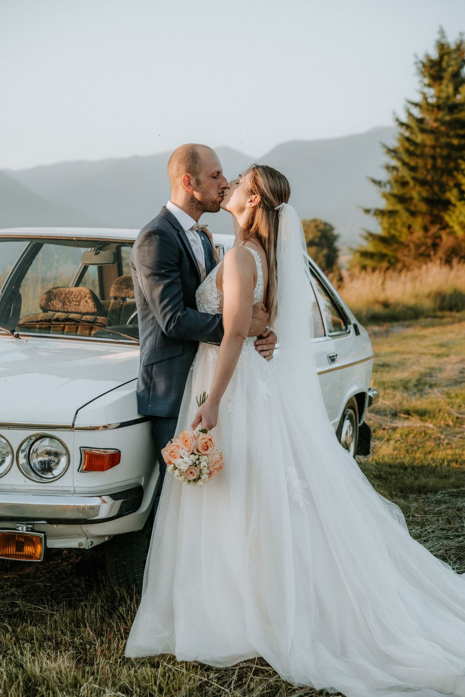 Newlyweds share a kiss beside a vintage white car in a scenic outdoor field at sunset.