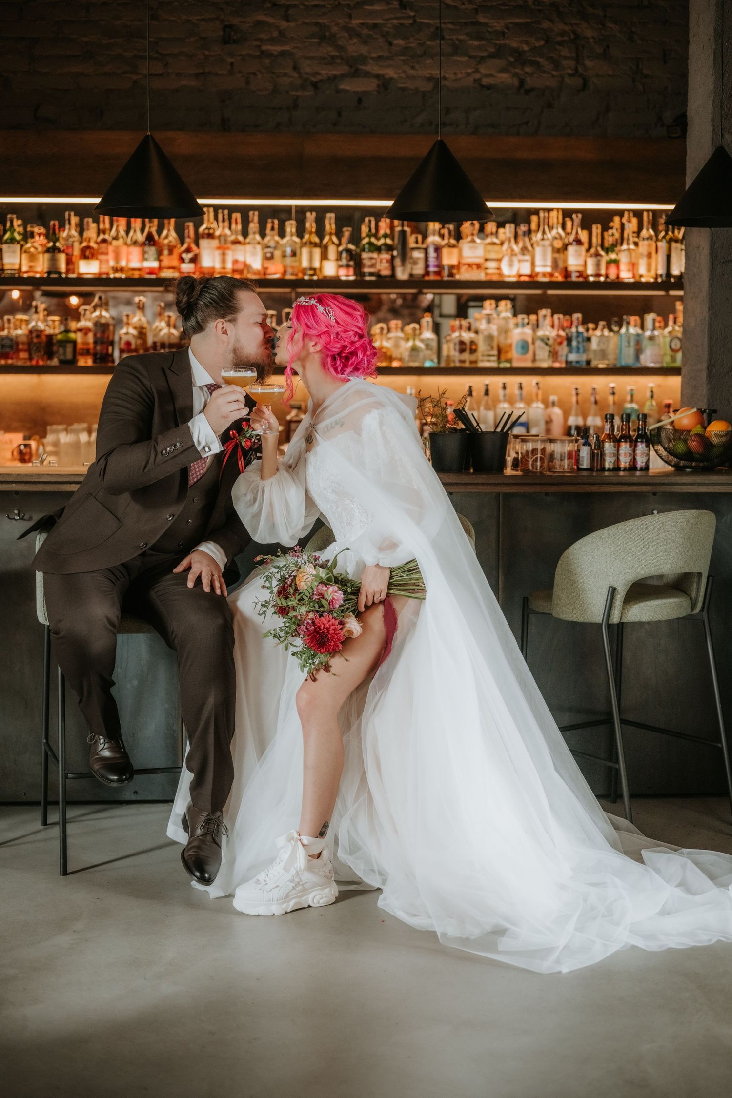 Bride with pink hair and groom share a kiss at a bar, she holds a red bouquet in a white gown with a long veil.