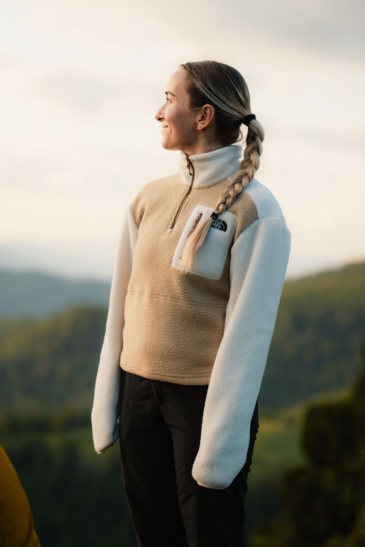 A woman in a two-tone North Face fleece pullover smiles while enjoying a scenic mountain view at dusk.