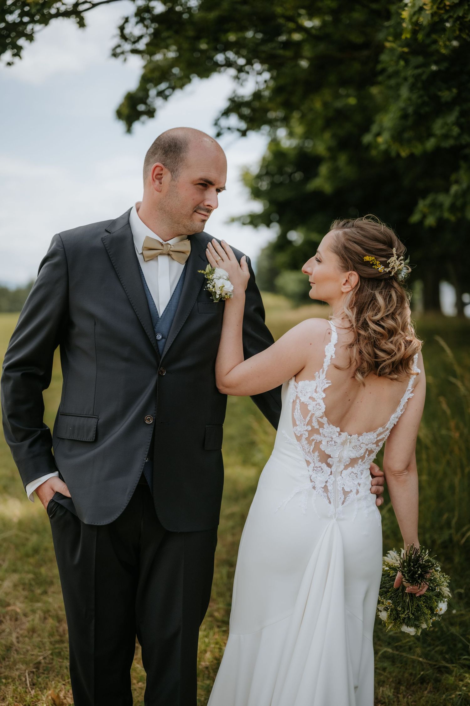 Bride and groom share a tender moment outdoors, she in lace-back white gown holding bouquet, he in dark suit with gold bow tie.