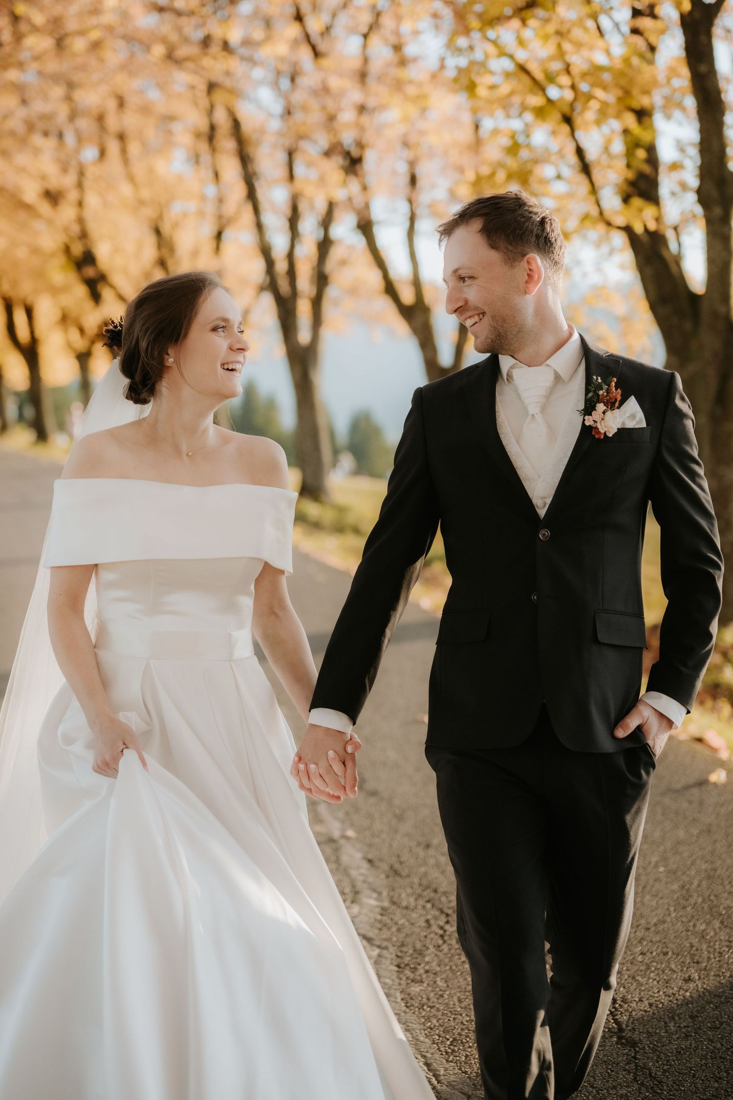 A smiling bride and groom walk hand in hand along a tree-lined path surrounded by golden autumn foliage.
