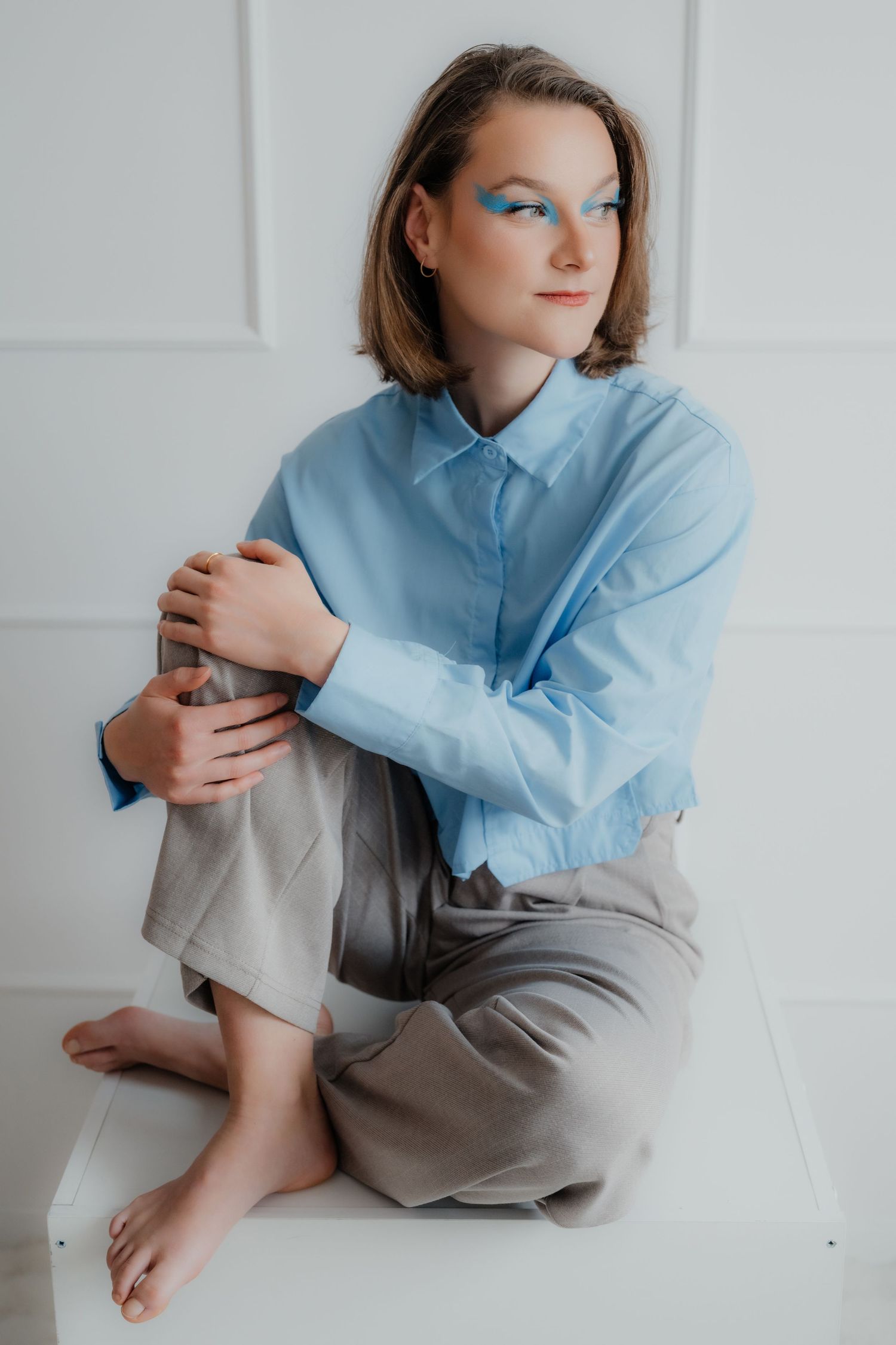 Woman with blue eye makeup in blue shirt sitting cross-legged on white cube.