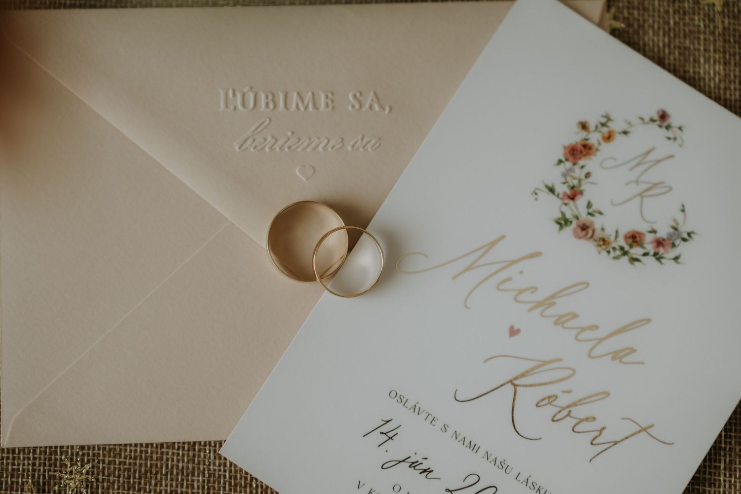 Wedding invitation with floral wreath, gold calligraphy names, two gold rings, and elegant envelope on burlap.