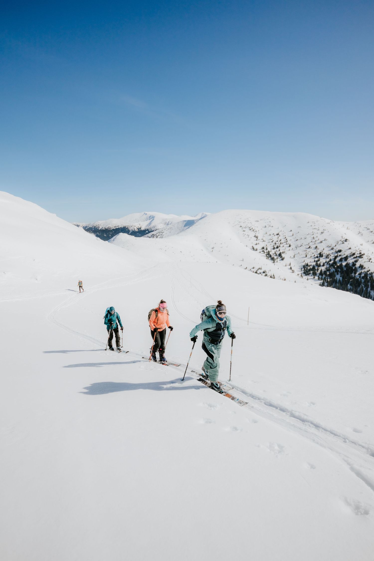 Three ski tourers ascending a wide snowy mountain slope with snow-covered peaks in the background.
