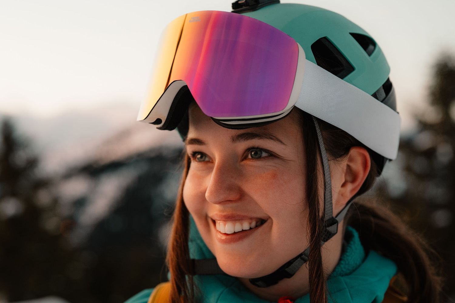Happy female skier with white helmet and iridescent pink ski goggles smiling in snowy mountain setting.