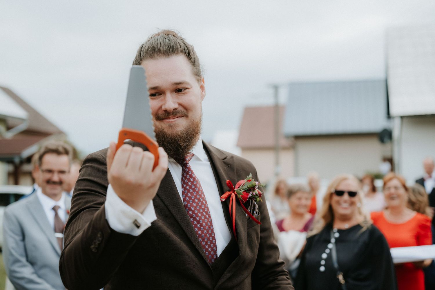 Bearded groom in brown suit holding a cleaver at an outdoor wedding ceremony with guests in background.