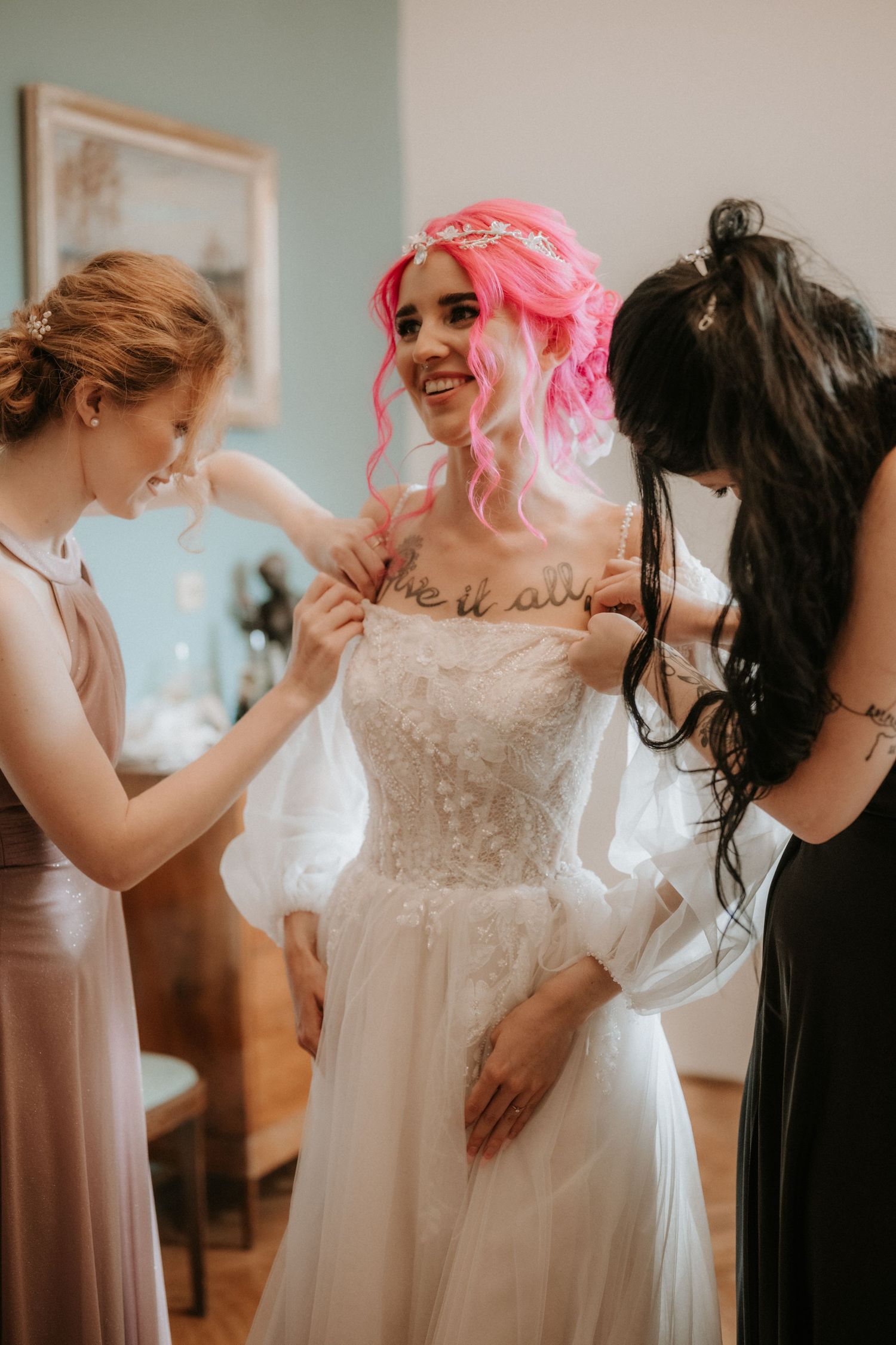 Bride with pink hair in white lace wedding gown assisted by two bridesmaids in elegant indoor setting.
