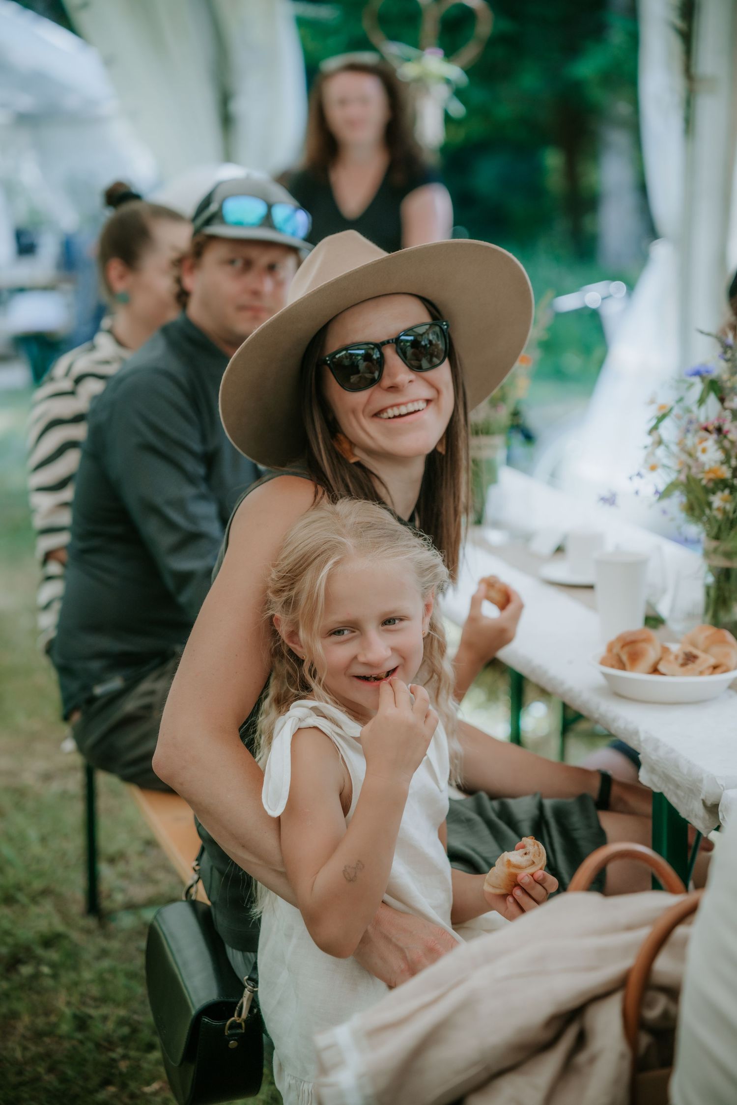 Woman in wide-brim hat and sunglasses smiling with young blonde girl at outdoor garden party event.
