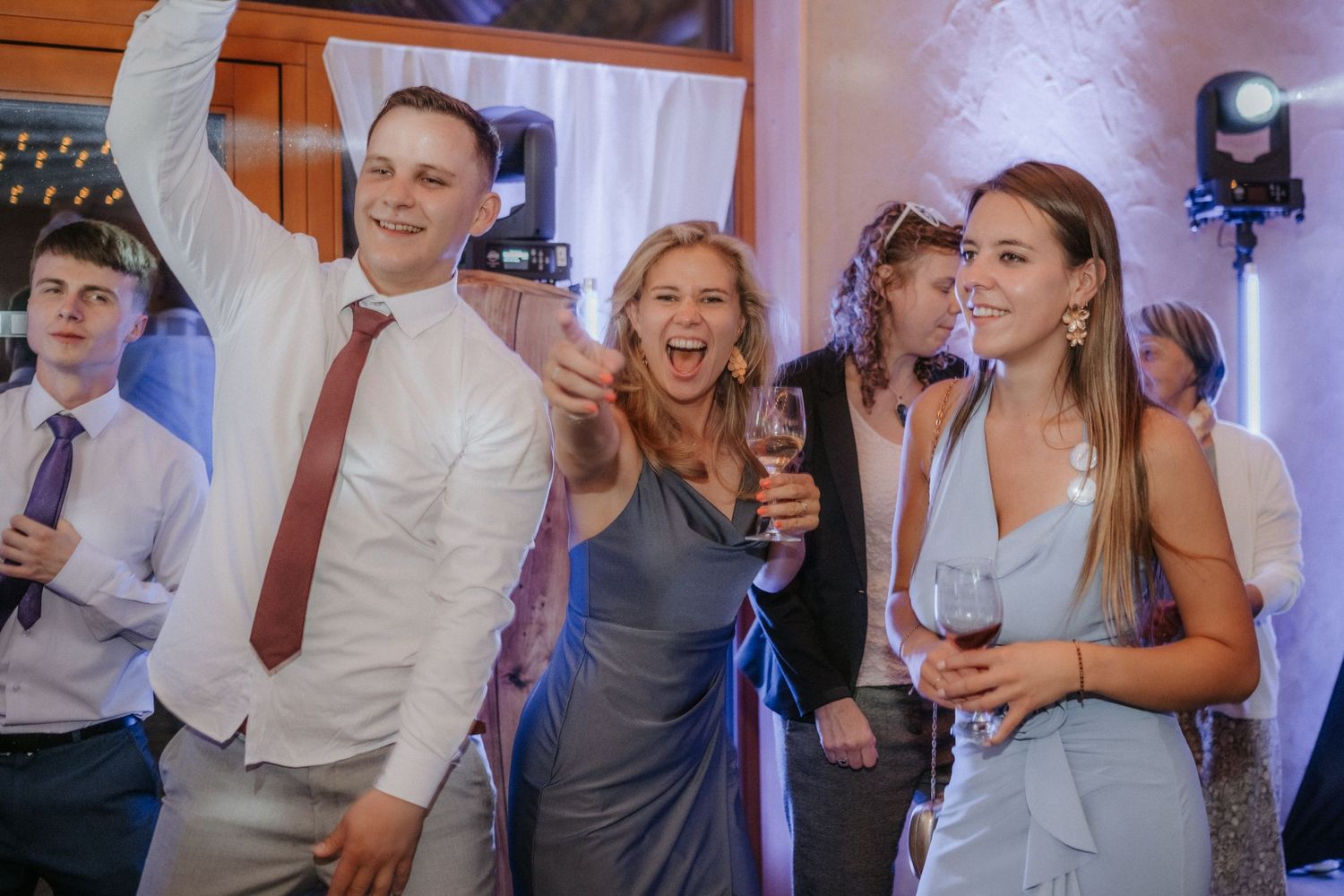 Guests celebrating at a wedding reception, dancing and holding wine glasses in a decorated venue.