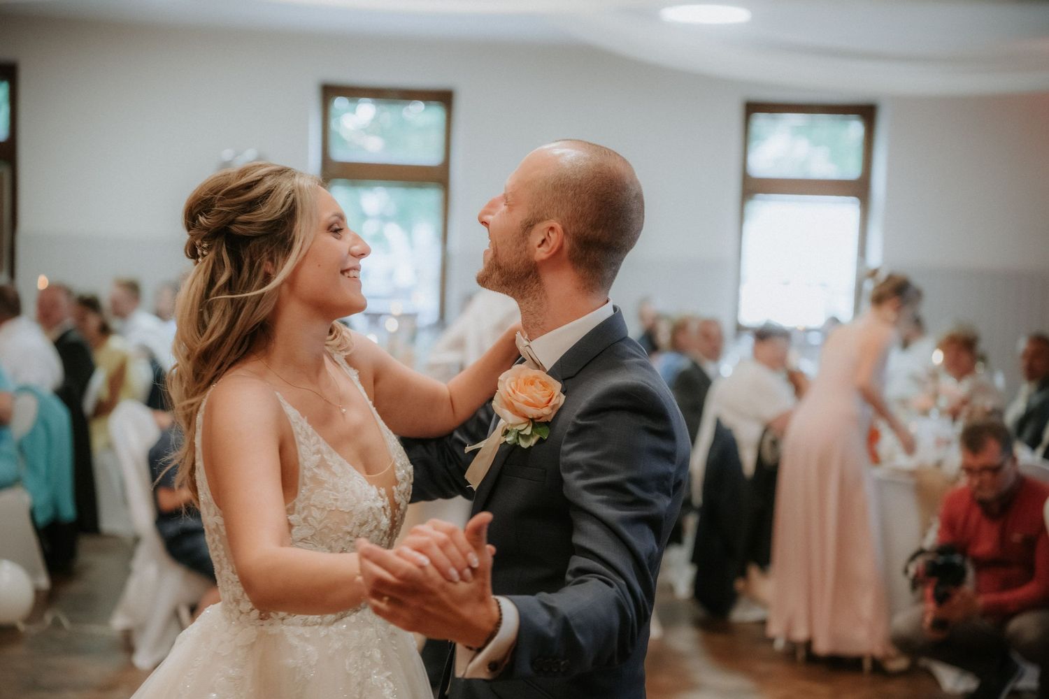 A bride and groom share their first dance, smiling at each other, surrounded by wedding guests in a bright reception hall.