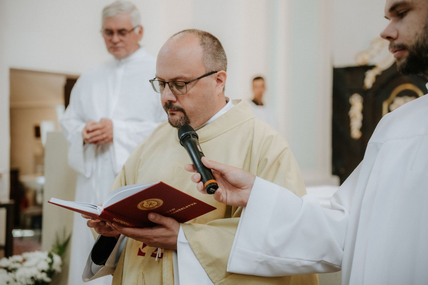 Catholic priest in white vestments reads from a red liturgical book during a church ceremony.