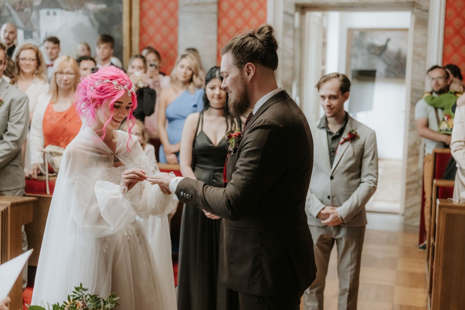 Bride with pink hair exchanges rings with groom in dark suit during wedding ceremony in a church.