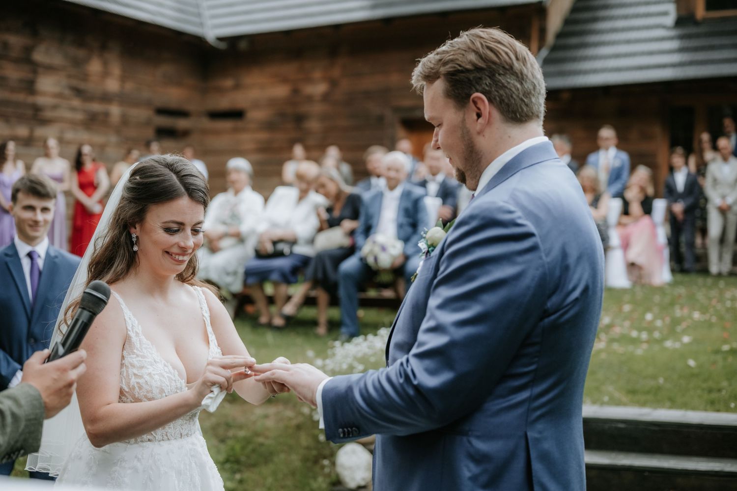 Bride and groom exchanging rings during outdoor wedding ceremony with guests seated in background.