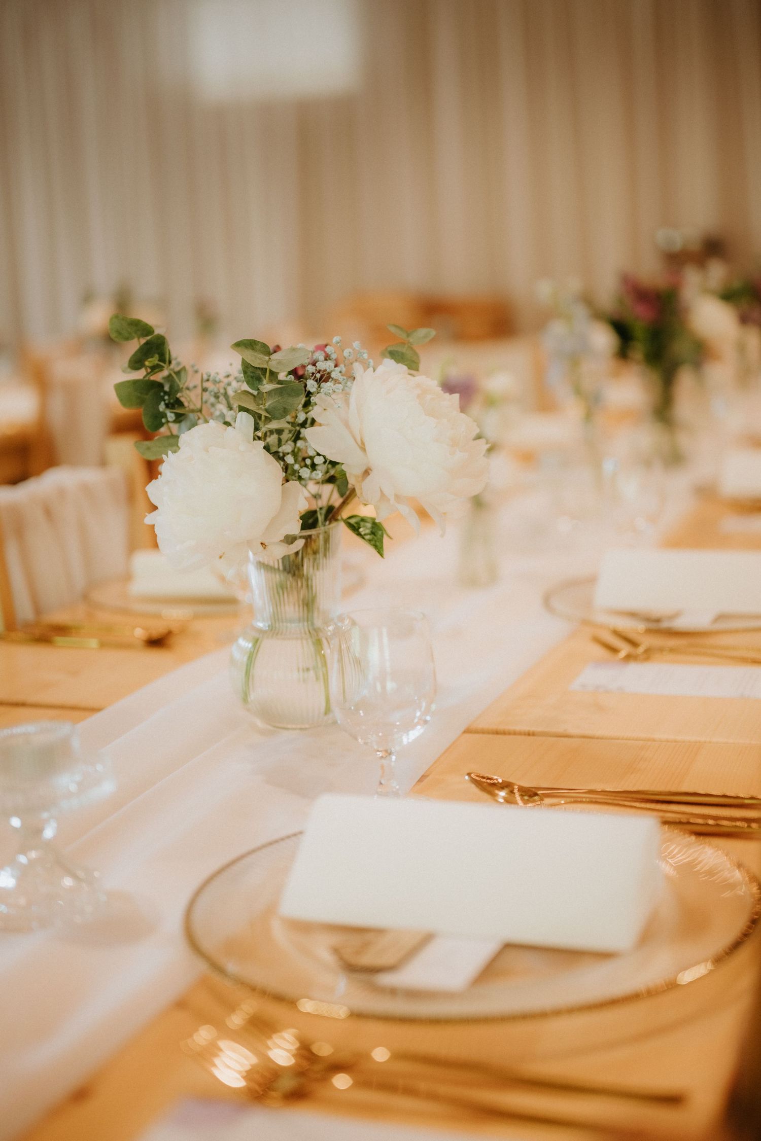 Elegant wedding reception table with white rose centerpieces, gold cutlery, glass charger plates, and a white table runner.