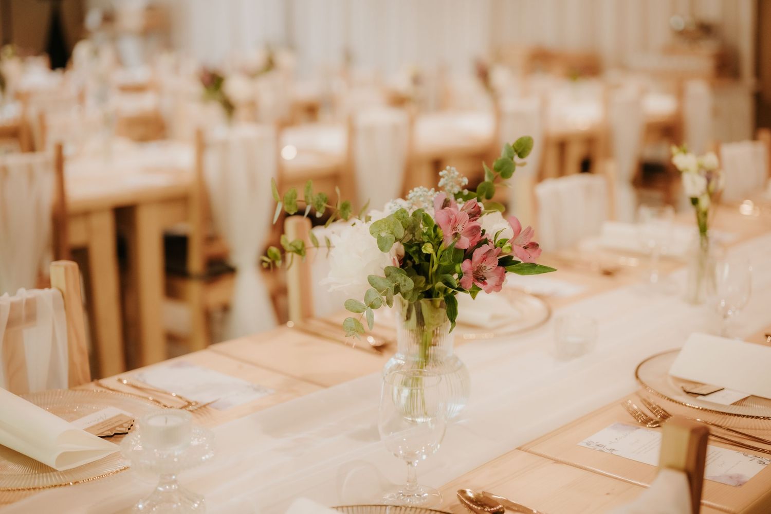 Elegant wedding reception table with floral centerpieces, white linens, and gold accents in a softly lit venue.