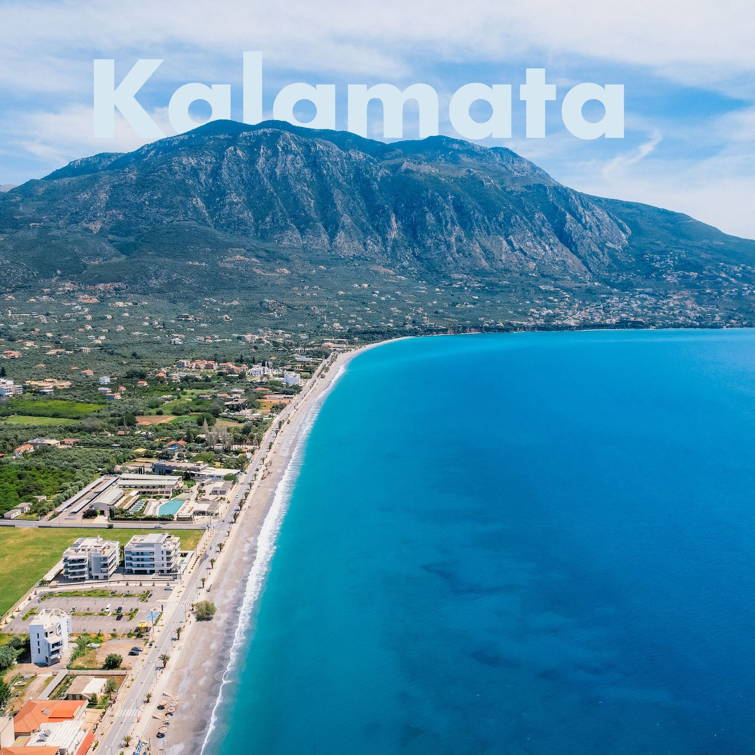 Aerial view of Kalamata, Greece, showing a long sandy beach, blue sea, and mountainous backdrop.