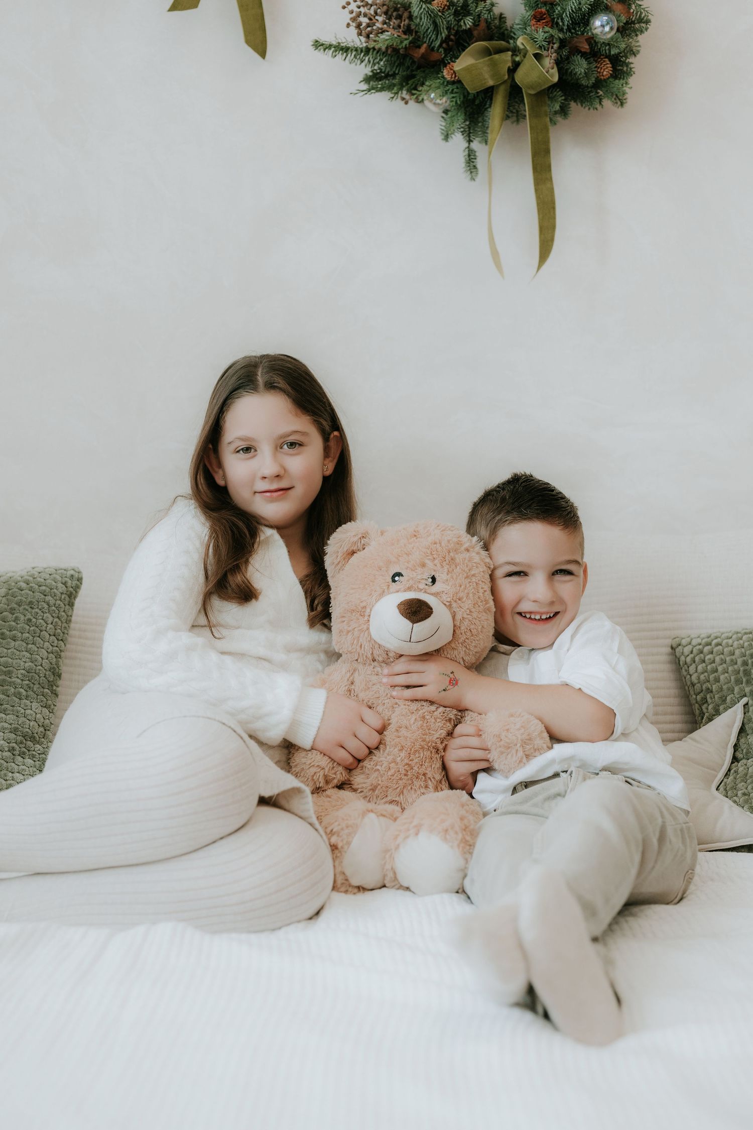 Two children in white pajamas sit on a bed hugging a teddy bear with a Christmas wreath on the wall behind them.