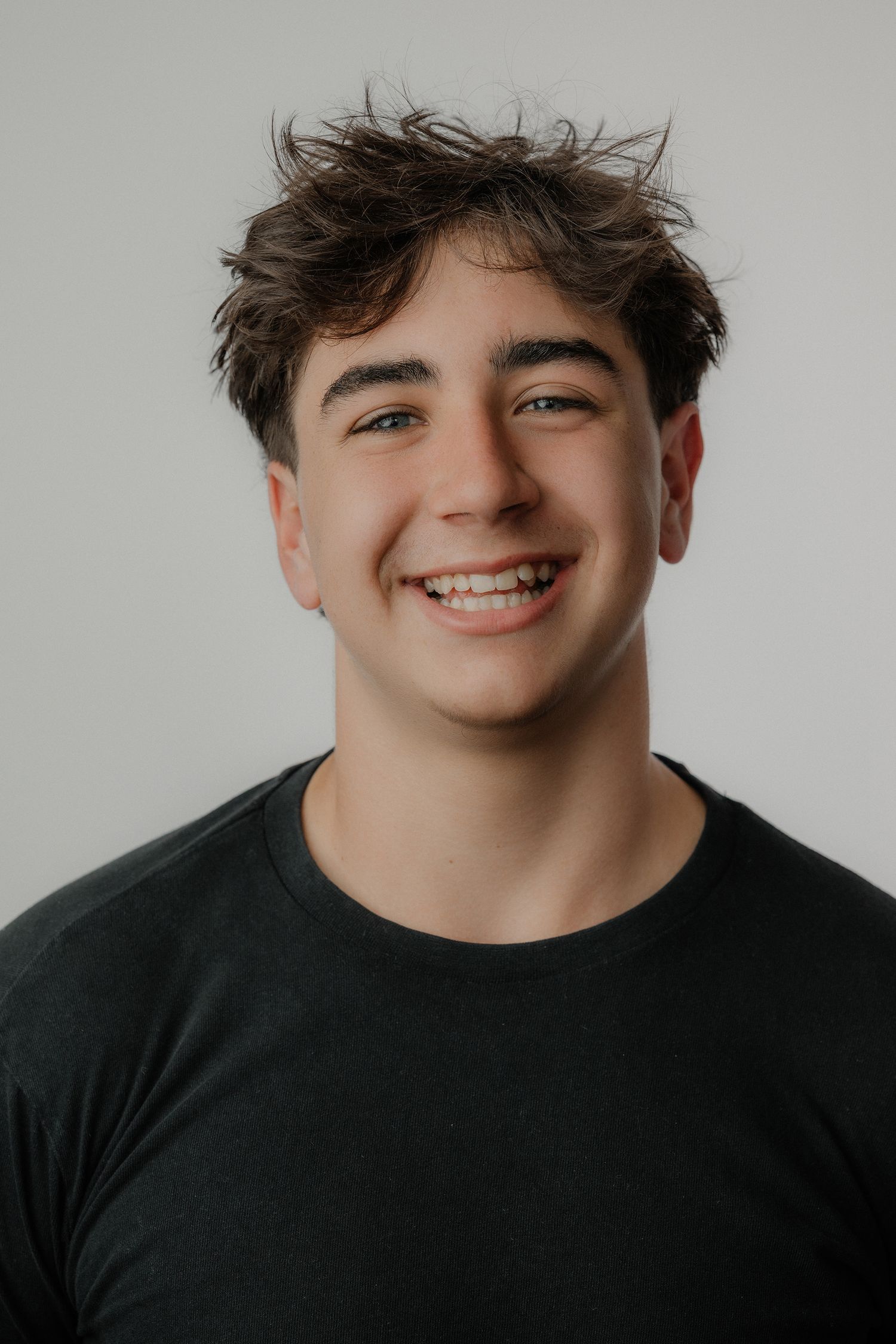 Smiling teenage boy with braces and dark hair wearing a black t-shirt against a white background.