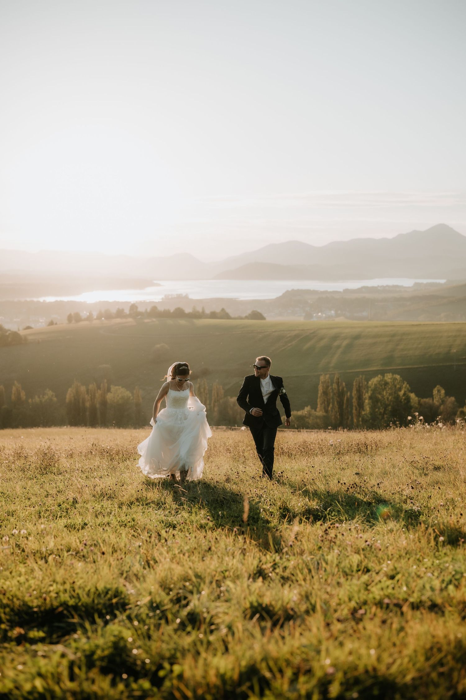 Bride and groom running through golden sunlit meadow with scenic mountain and lake backdrop on wedding day.