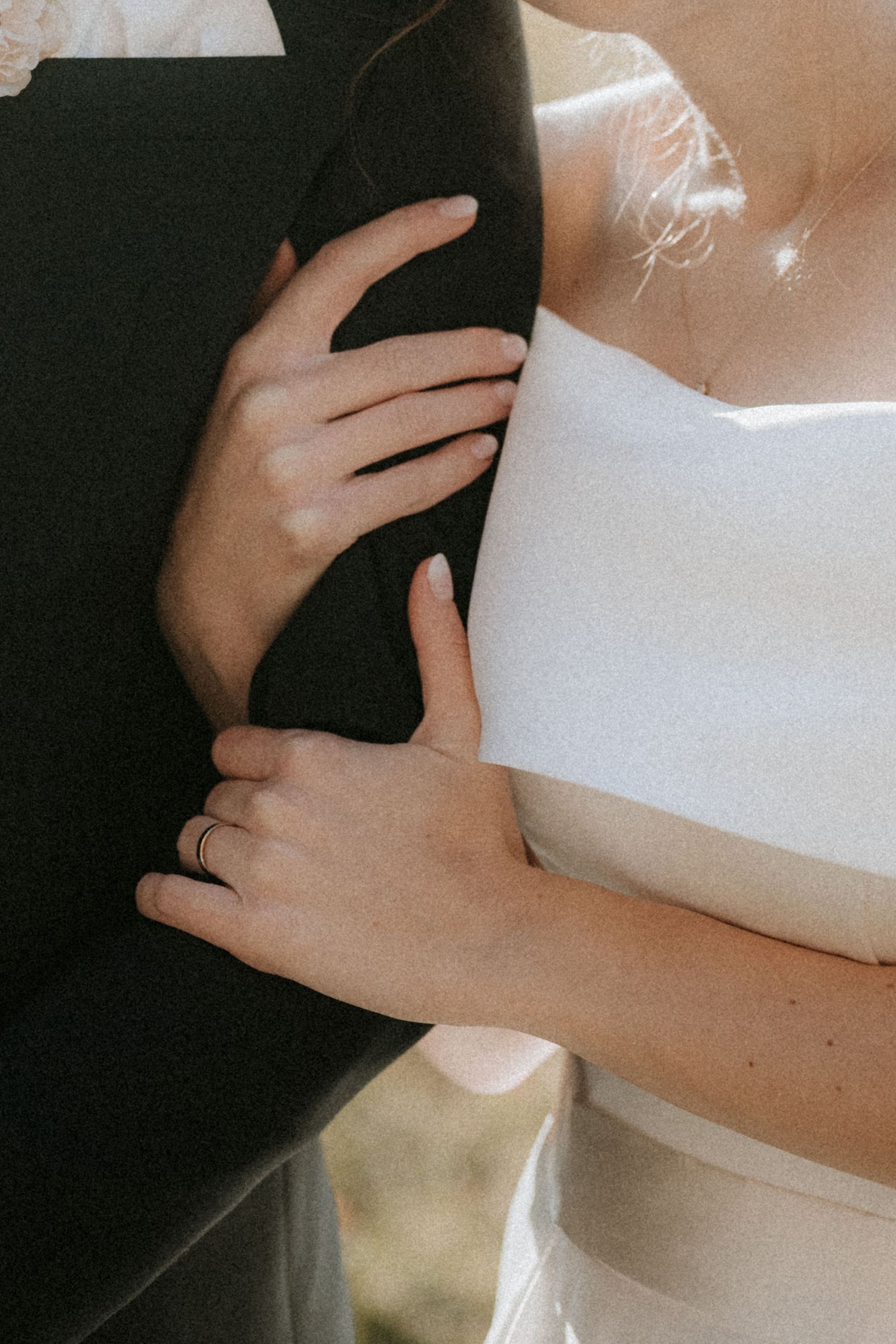 Close-up of bride and groom embracing, showing wedding rings on hands, white bridal gown and dark suit.