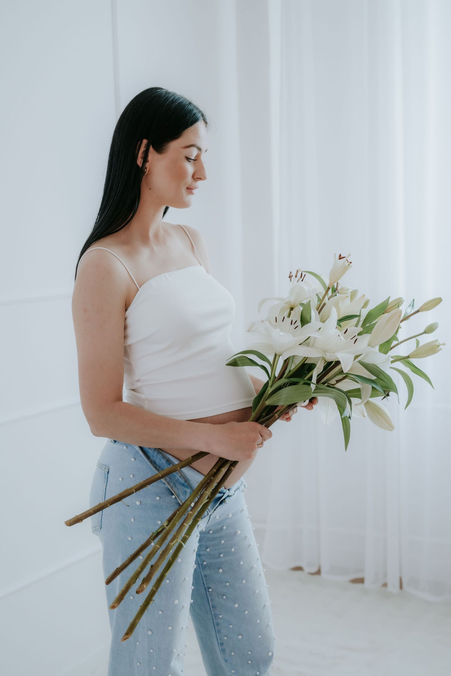 Pregnant woman in white crop top holding elegant white lily bouquet, side profile view.