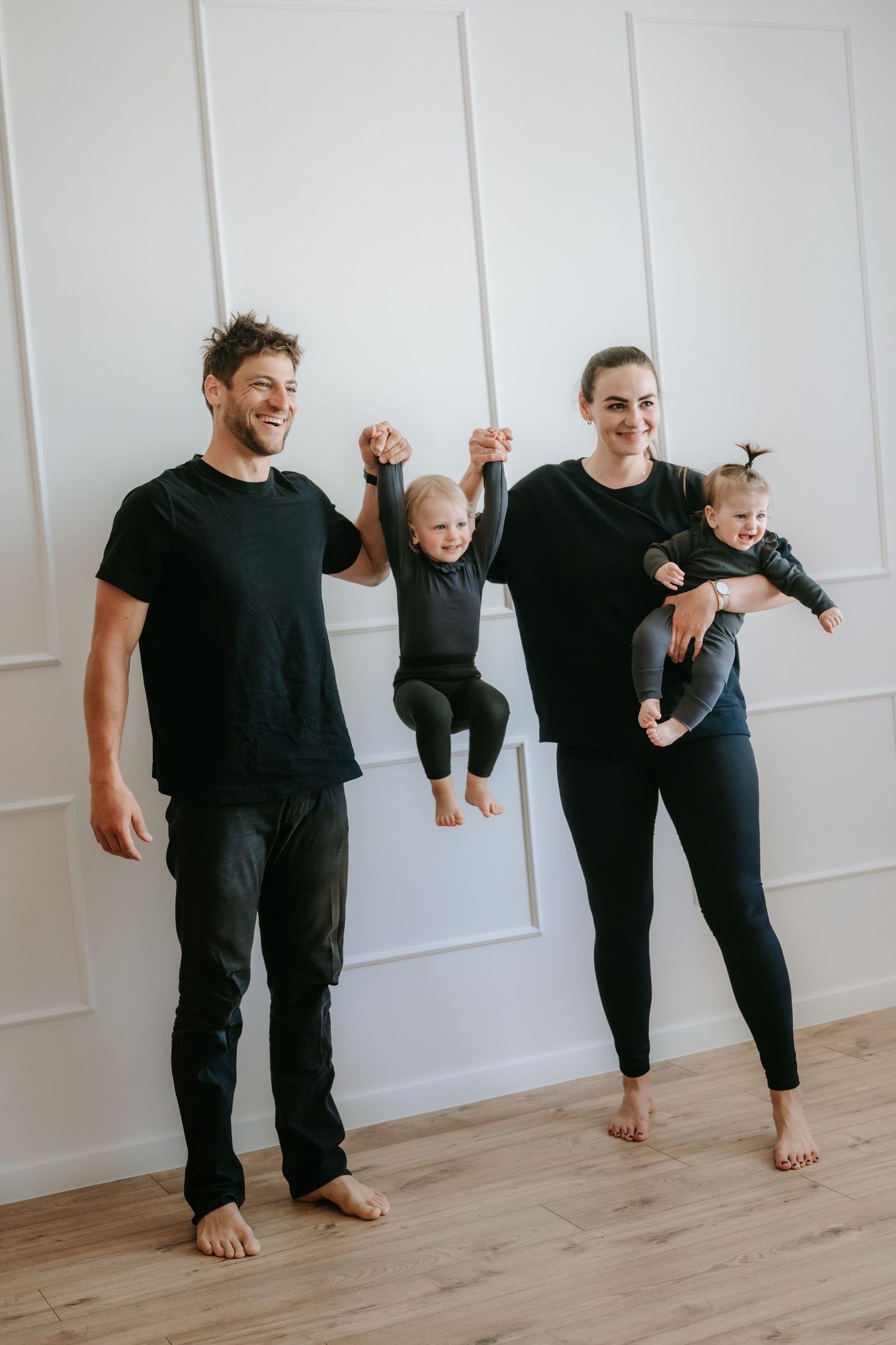 Happy family in matching black outfits, father lifting toddler while mother holds baby against white wall.