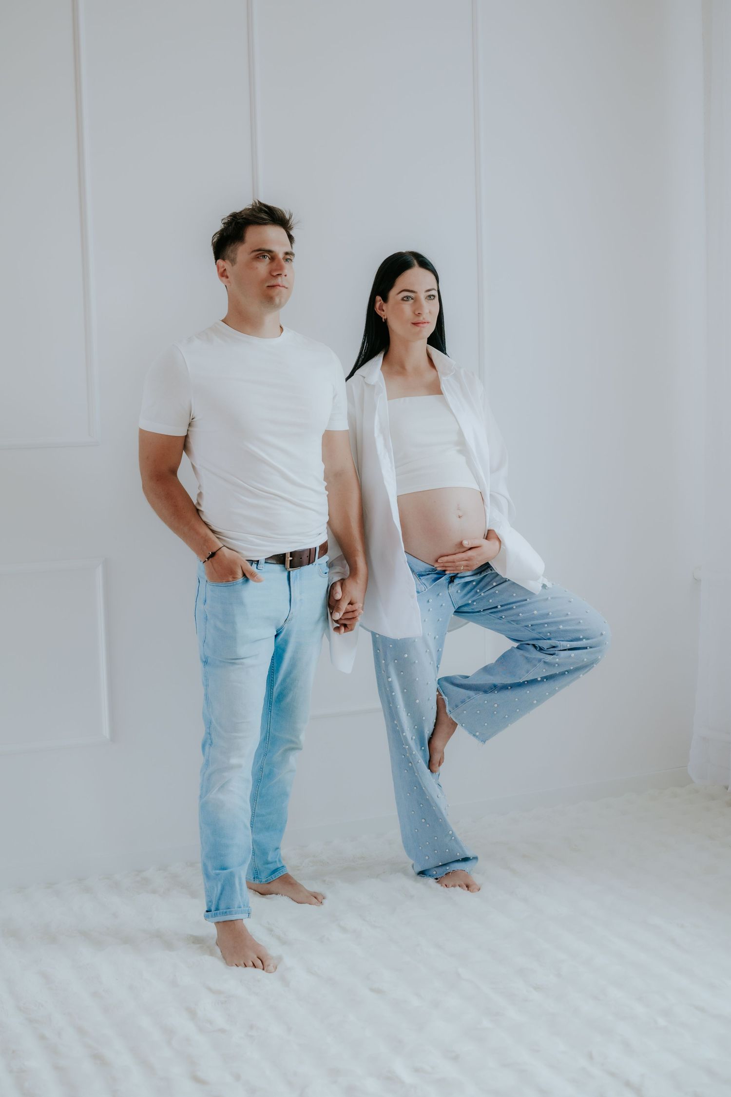 Pregnant woman in white crop top and jeans stands with partner in bright white studio setting.