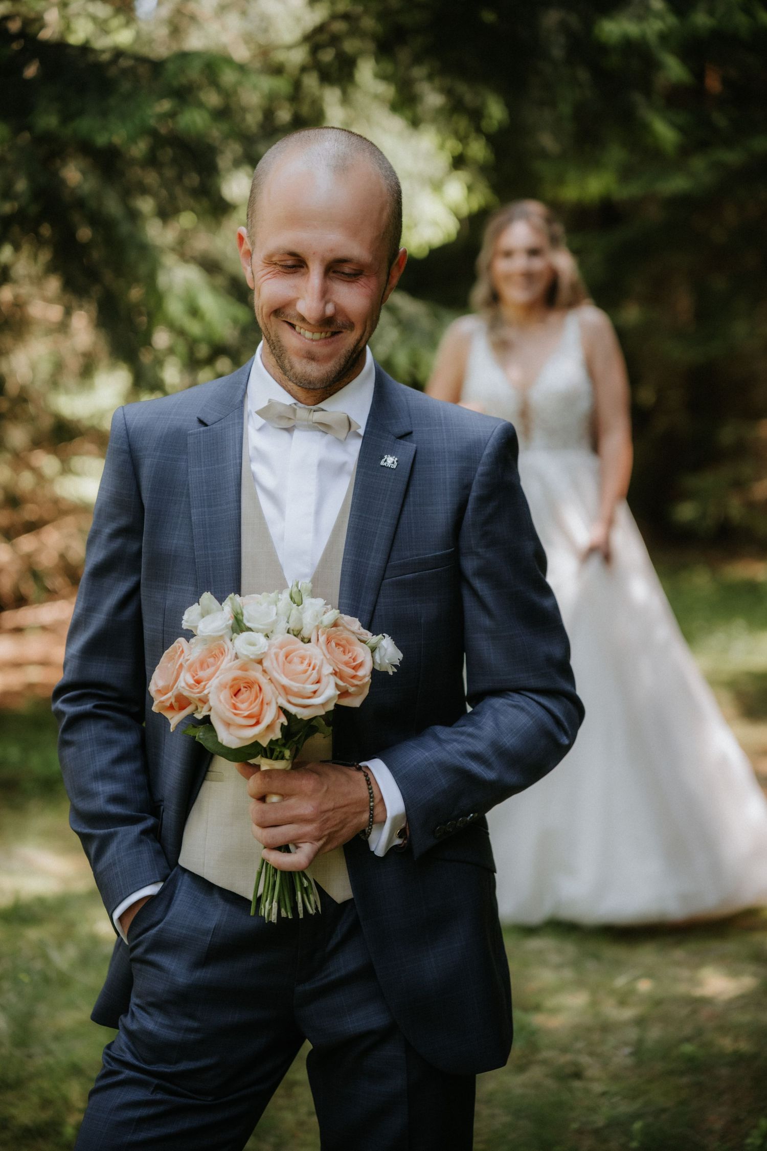 Smiling groom in navy suit holding pink rose bouquet with bride in white gown standing in background outdoors.