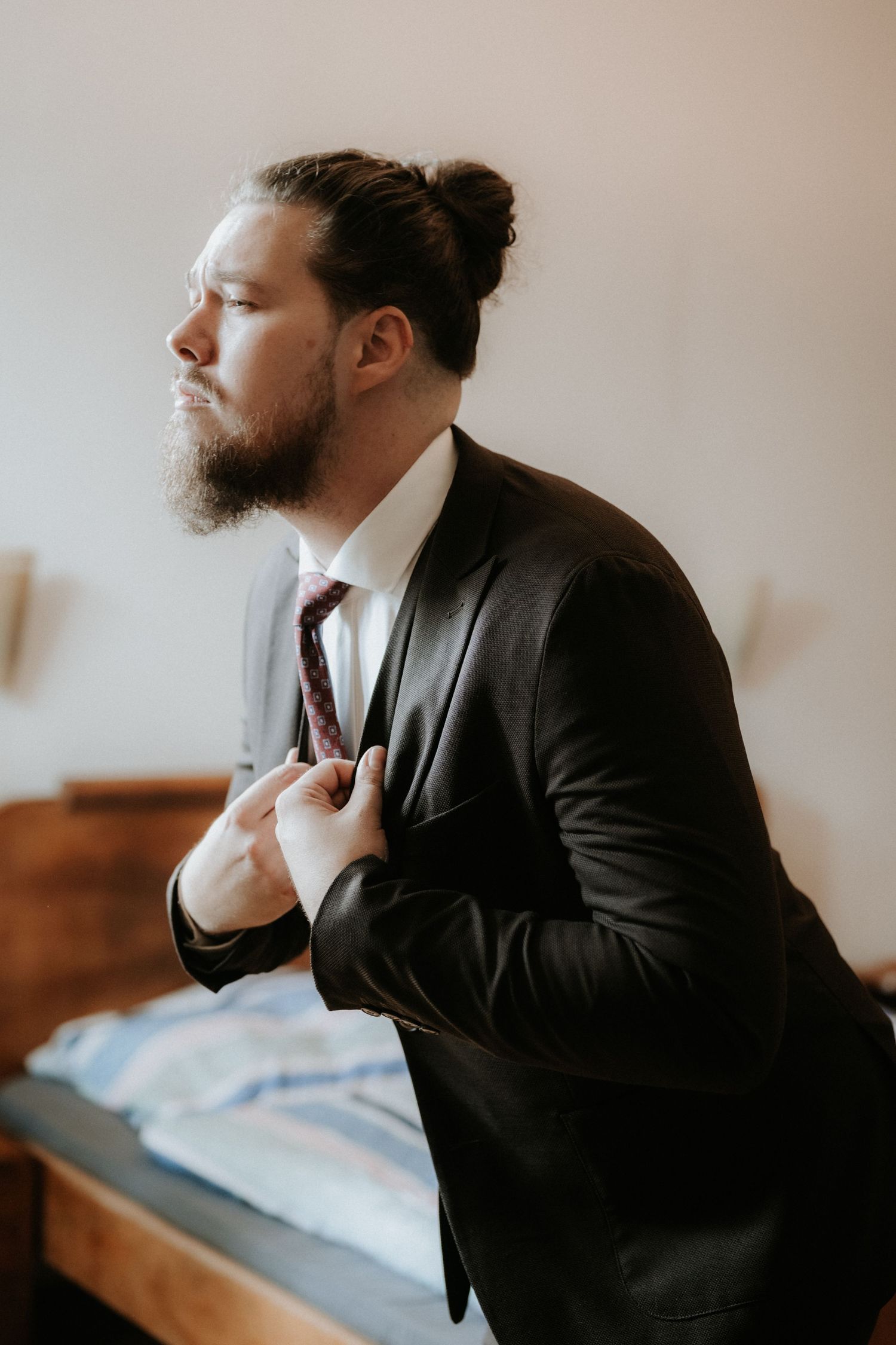 Bearded man in black suit adjusting burgundy tie with hair in bun, getting dressed in elegant room.
