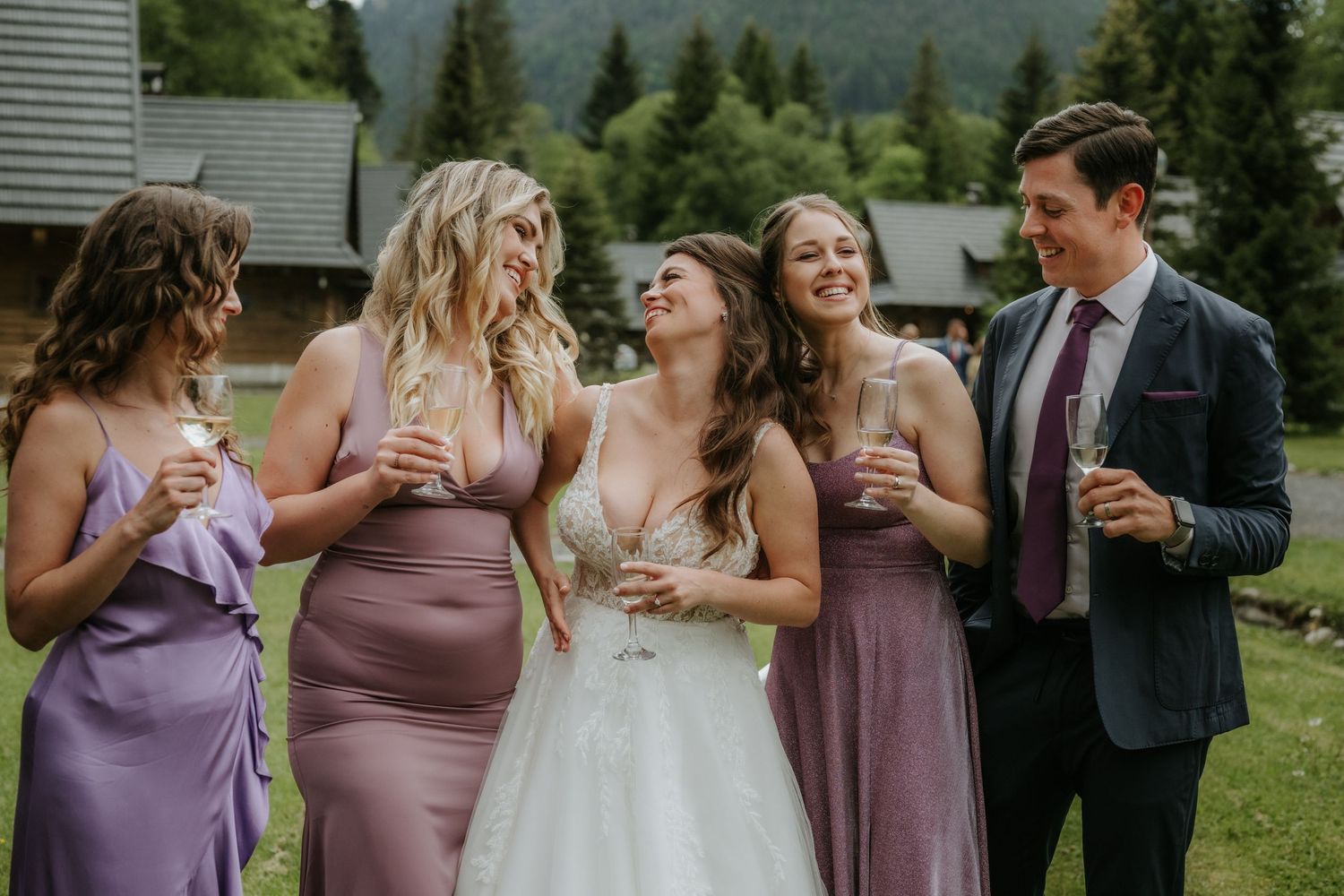 Bride in white gown laughing with bridesmaids in purple dresses and groomsman holding champagne outdoors.