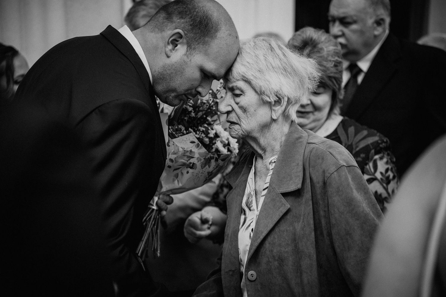 Black and white photo of a man in a suit kissing an elderly woman on the cheek at a formal gathering.