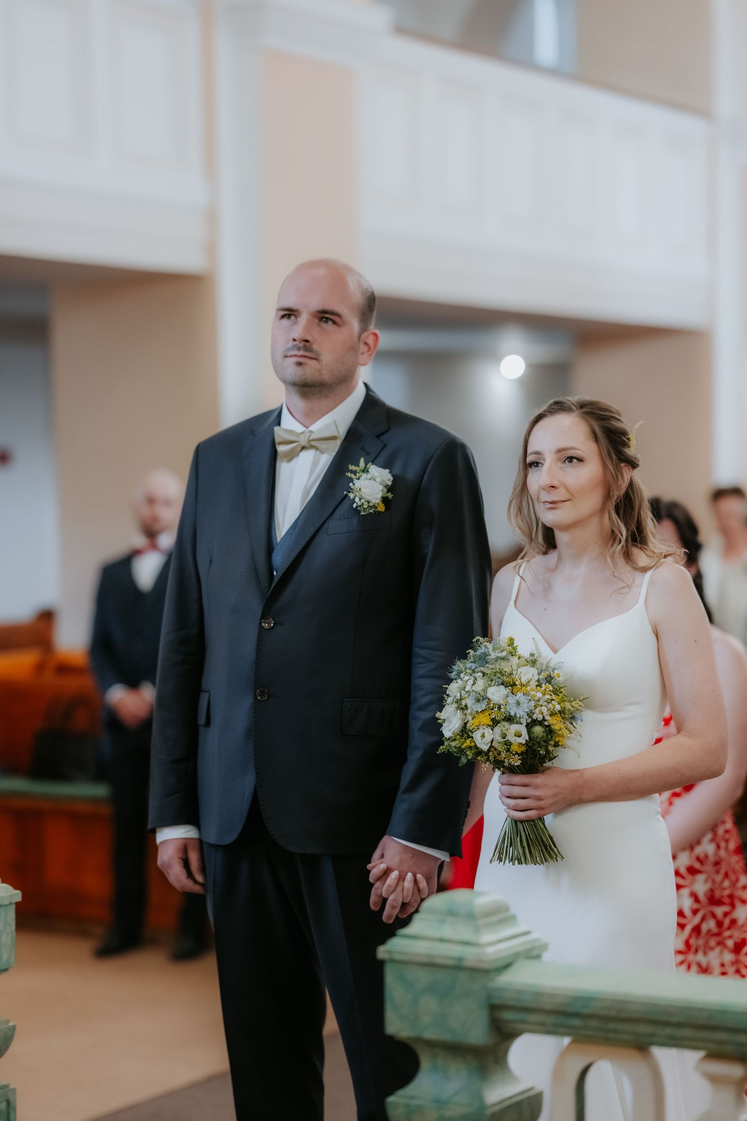 Bride and groom holding hands at wedding ceremony, bride in white dress holding wildflower bouquet, groom in navy suit.