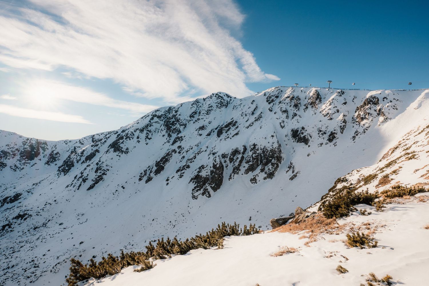 Snow-covered mountain peak under a blue sky with wispy clouds, featuring rocky terrain and sparse alpine vegetation in winter.