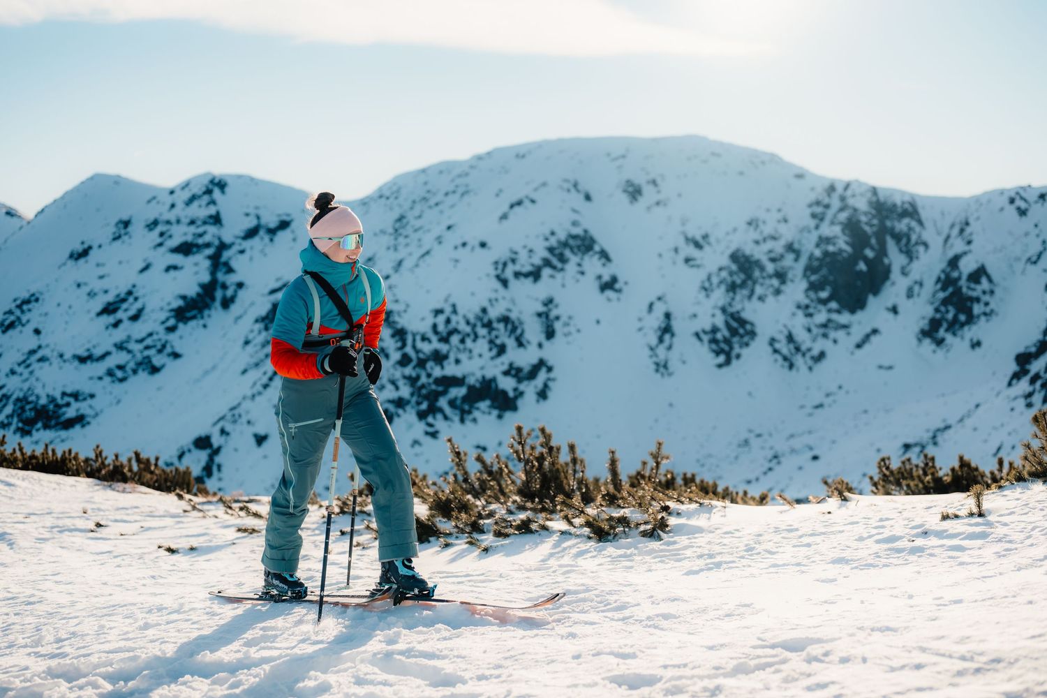 Woman on skis pausing on a snowy mountain ridge, wearing teal jacket and white hat on a sunny winter day.