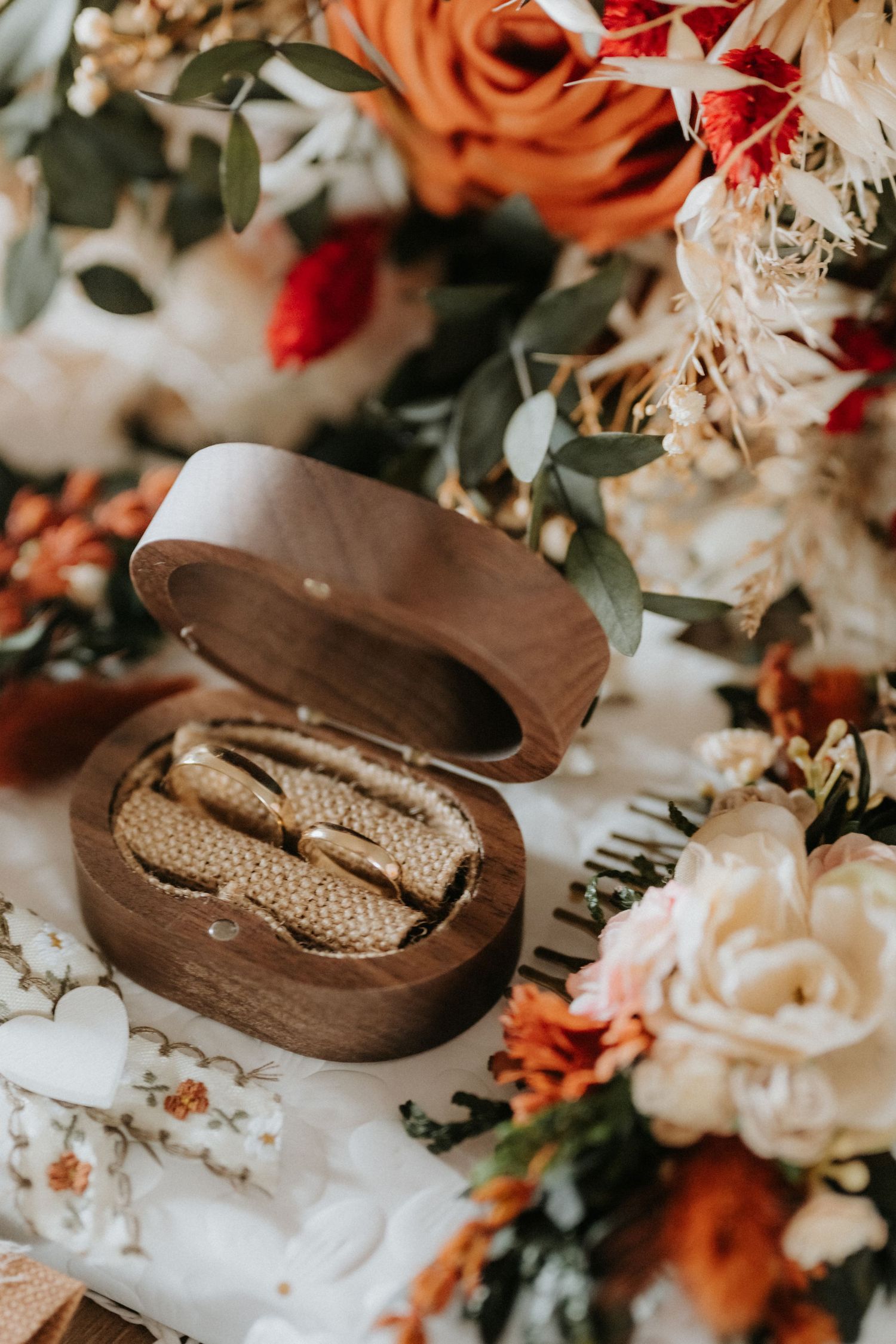 Wooden ring box with gold wedding bands surrounded by autumn floral arrangement featuring orange, cream, and red blooms.