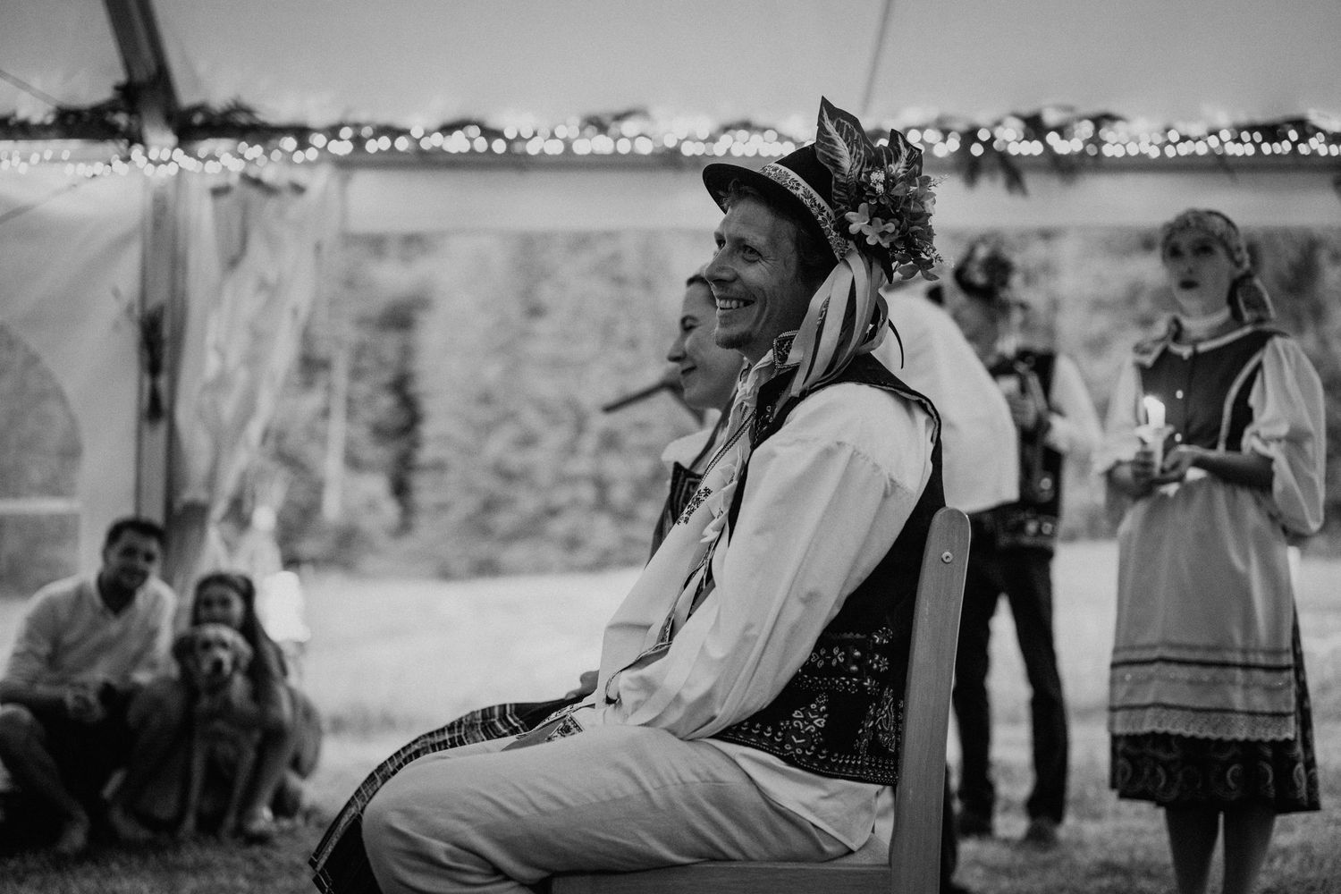 Black and white photo of a man in folk costume with floral crown seated outdoors at a traditional celebration.