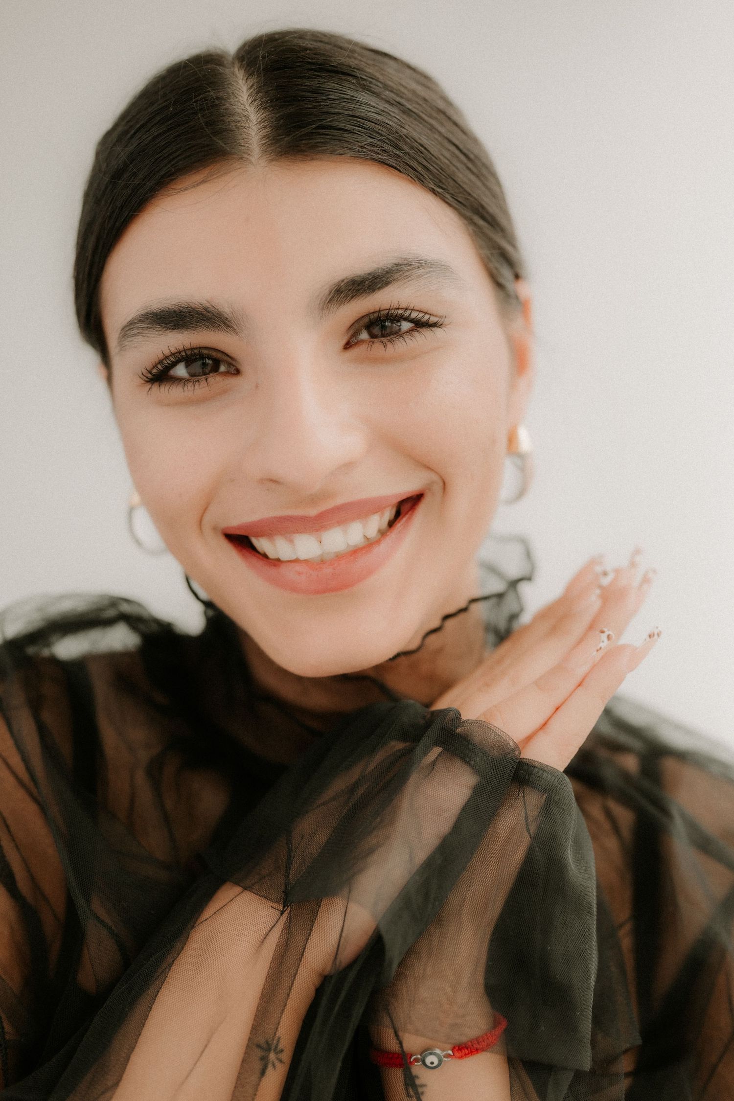 Smiling woman in black sheer top with decorative nails and red bracelet on white background.
