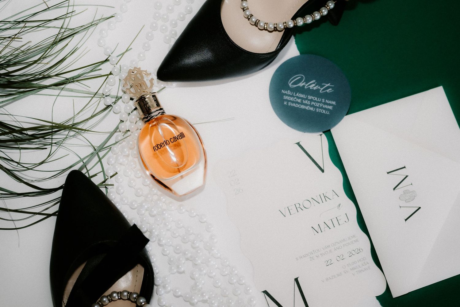 Flat lay of black heels, amber perfume bottle, pearl necklace, and wedding invitation on green backdrop.