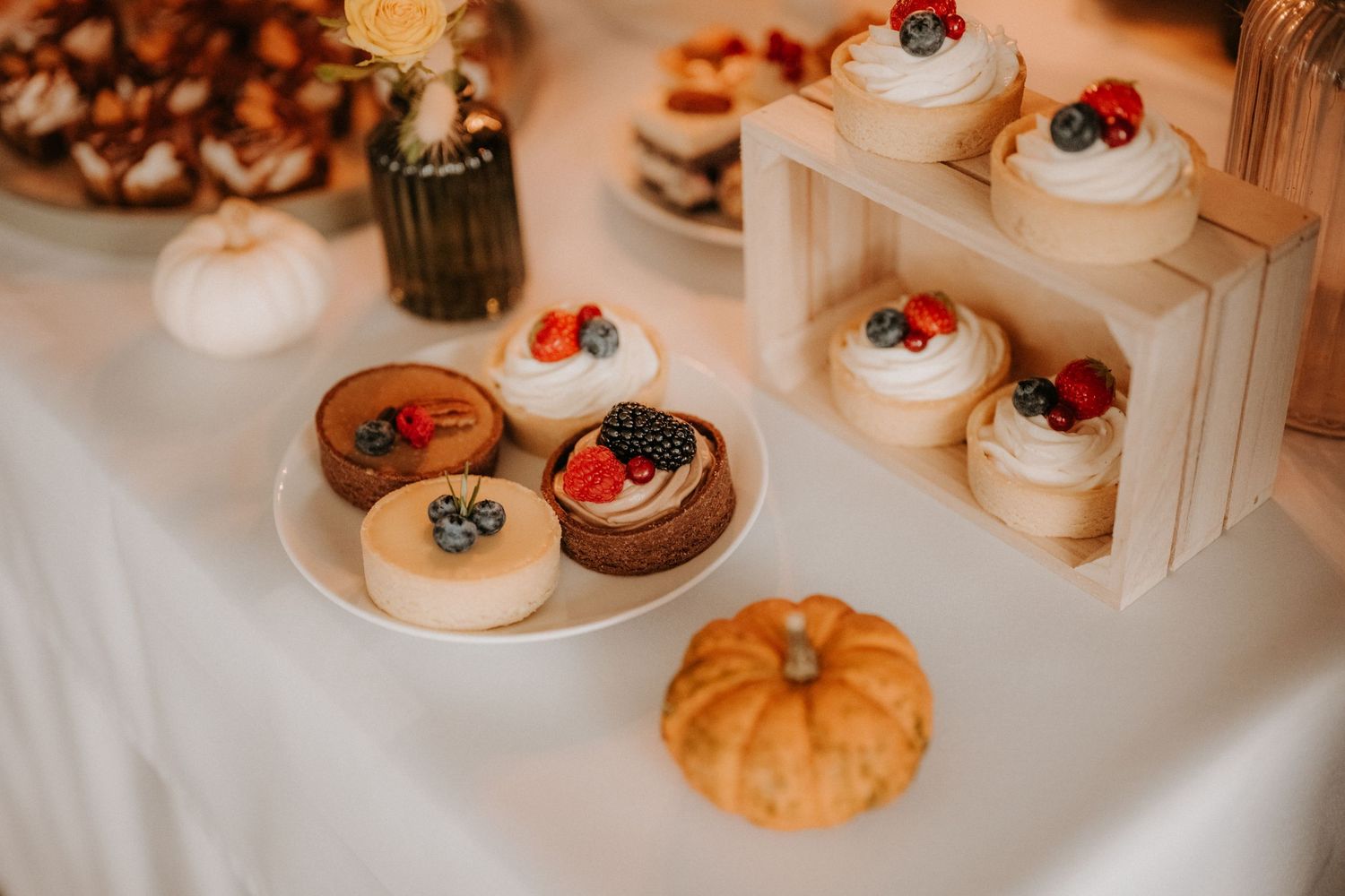 Elegant dessert table with cupcakes, tarts topped with fresh berries, and a small pumpkin at a fall wedding reception.