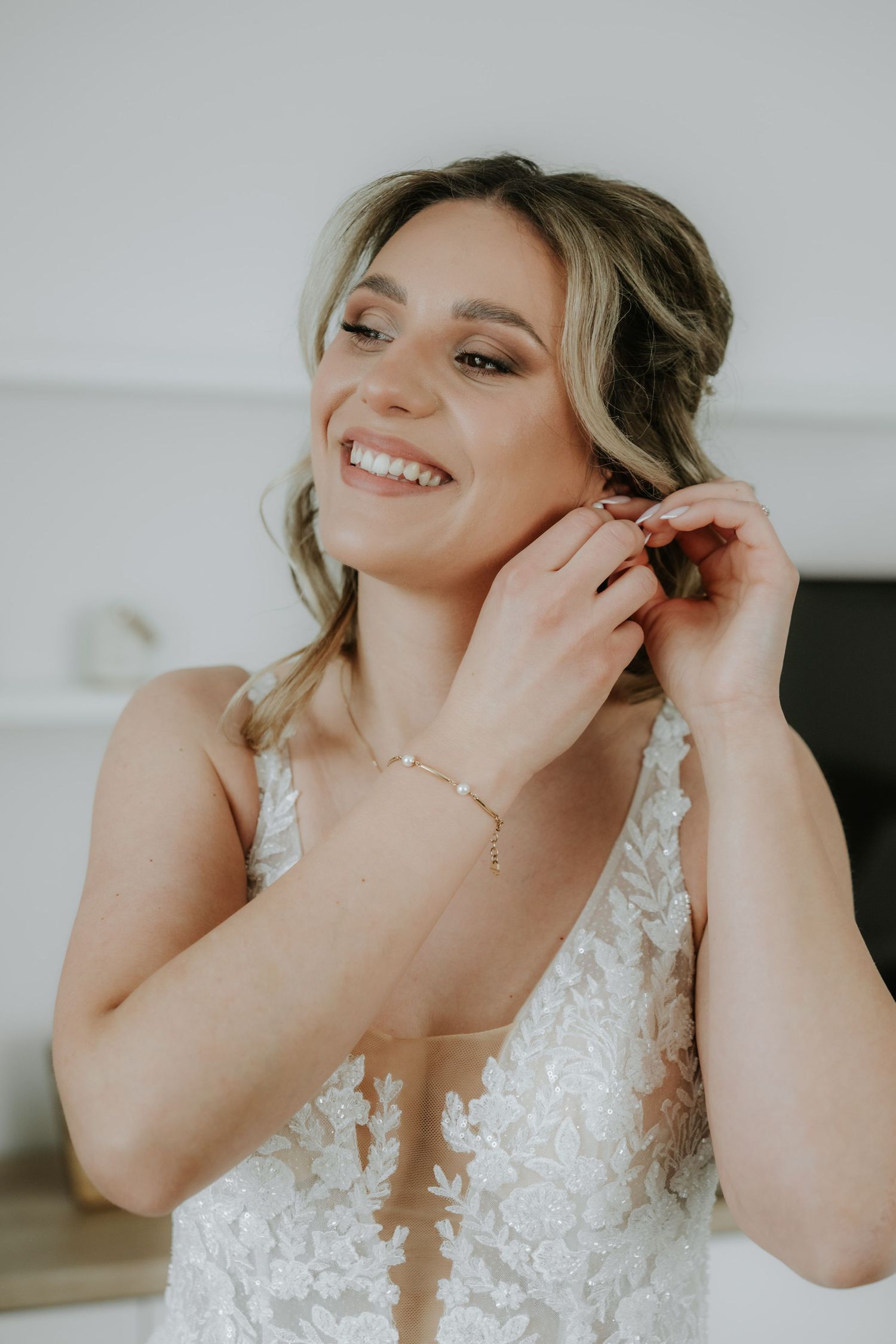 Smiling bride in lace wedding dress putting on earrings, wearing pearl bracelet and necklace.
