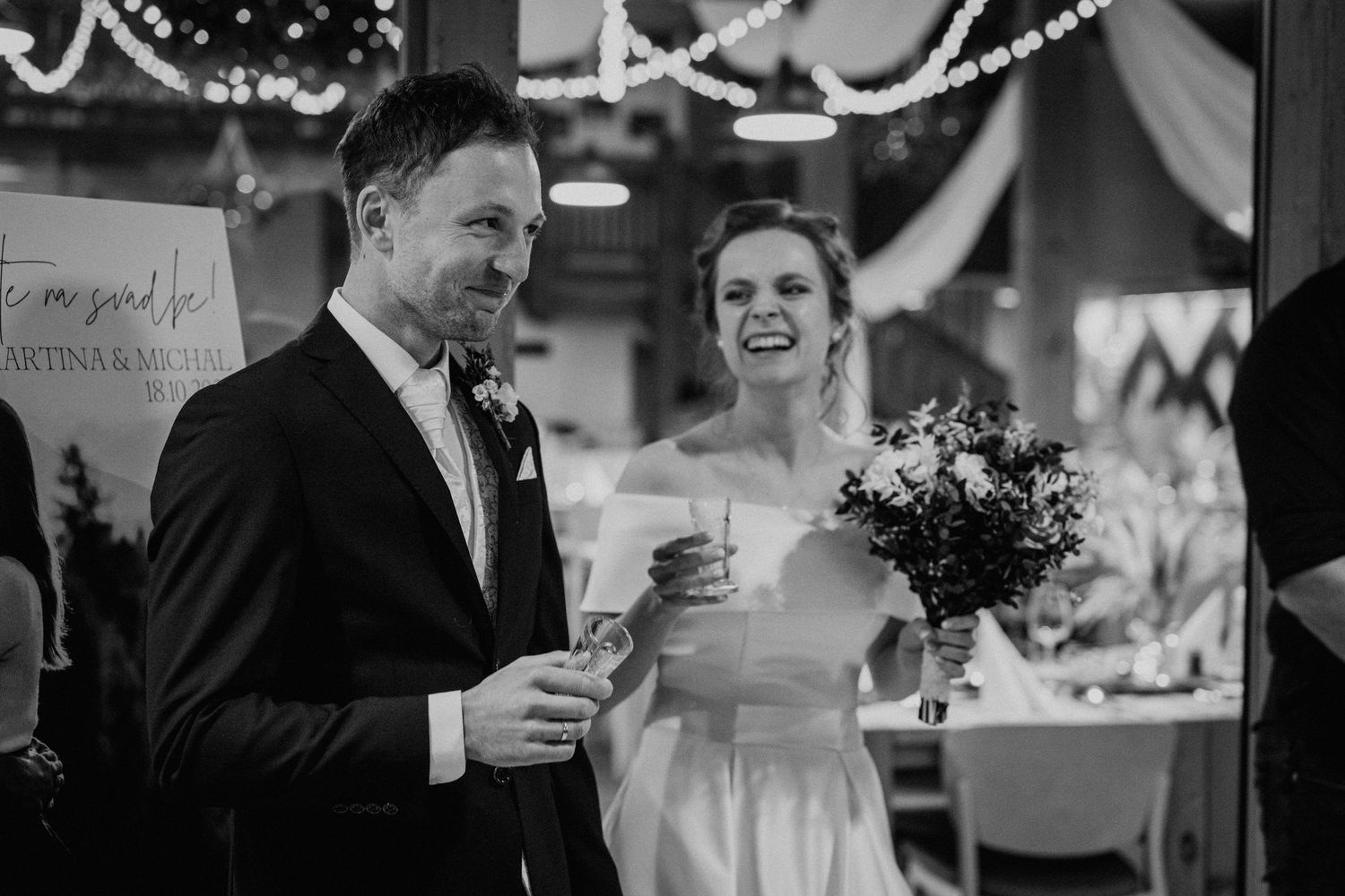Black and white photo of a smiling bride holding a bouquet and groom with a drink at a wedding reception.