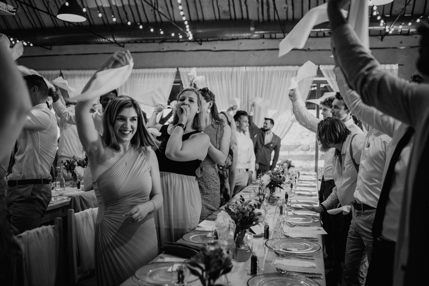 Black and white wedding reception photo with guests celebrating, waving papers, smiling along a decorated banquet table.