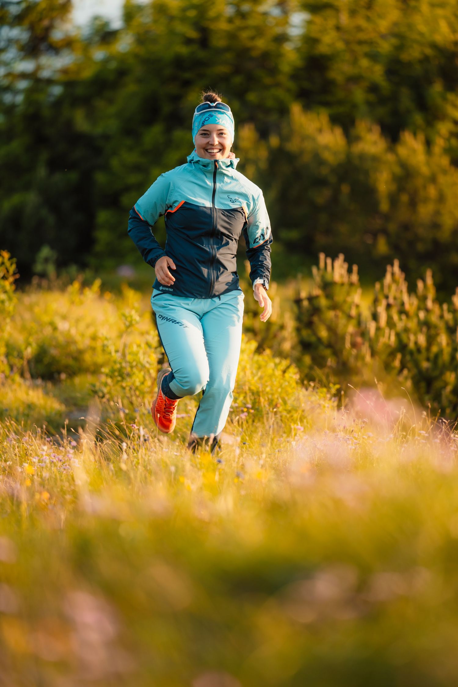 A smiling woman in teal athletic wear jogs through a vibrant sunlit meadow surrounded by green shrubs.