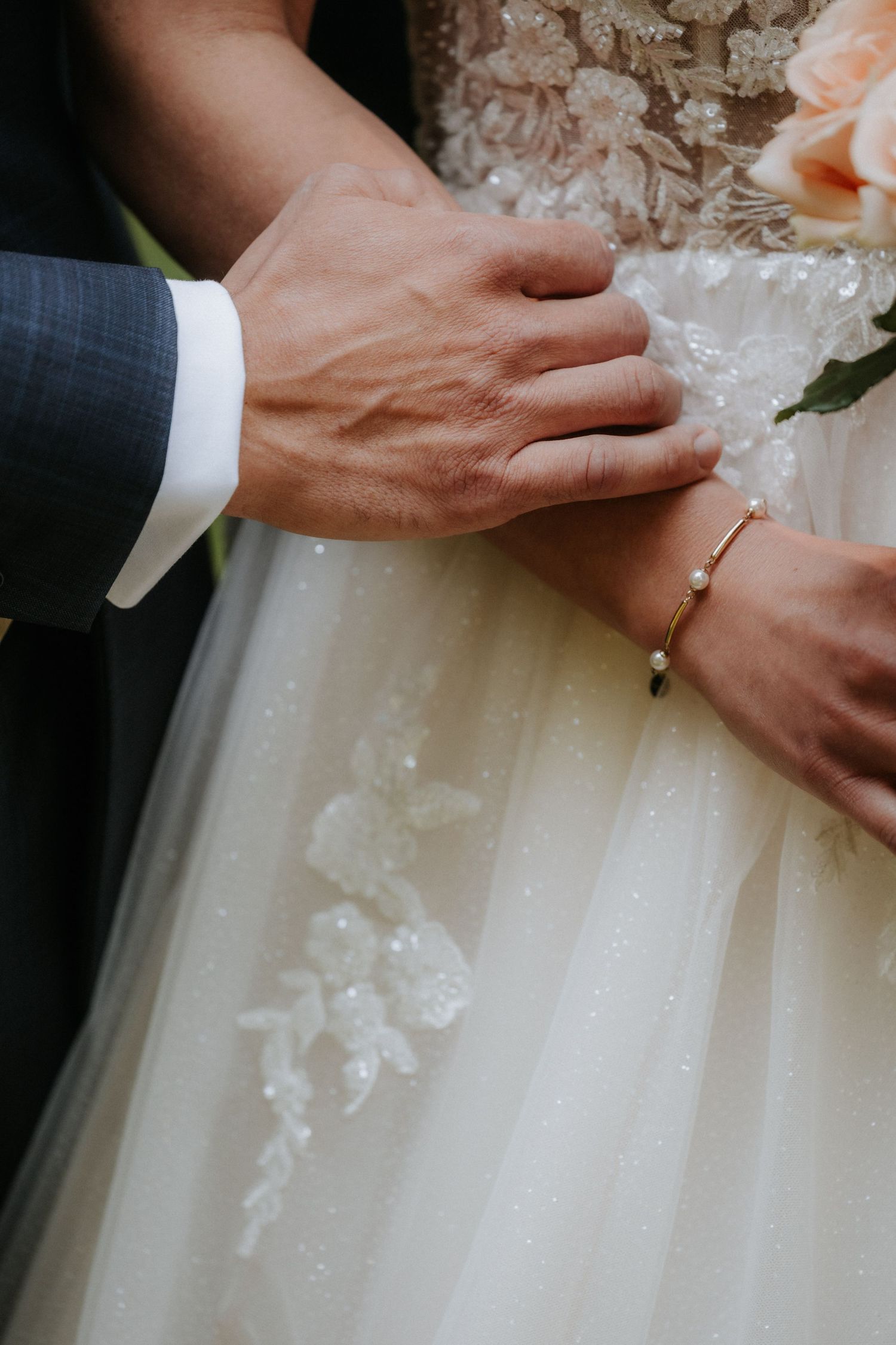 Close-up of bride and groom hands touching, bride wearing gold bracelet and lace wedding dress.